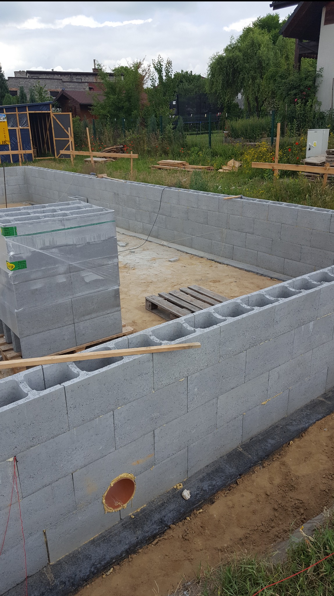 Wzniesione ściany fundamentowe z bloczków betonowych, z widoczną izolacją przeciwwilgociową i otworem instalacyjnym. Na palecie bloczki do dalszej budowy.