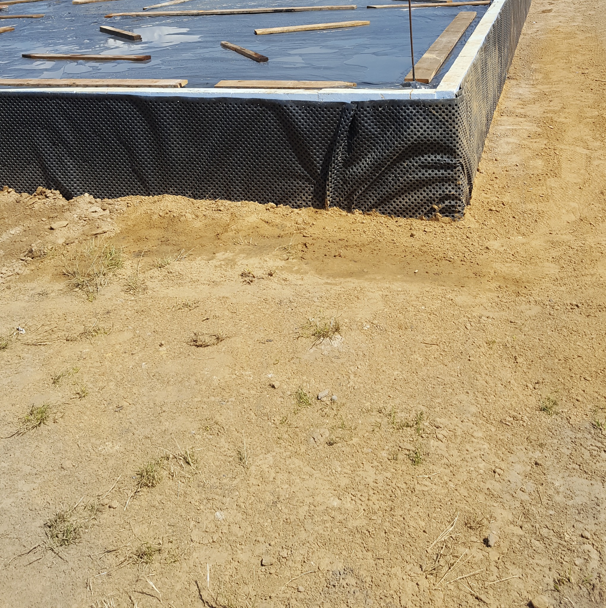 Zabezpieczone fundamenty z izolacją pionową i folią hydroizolacyjną. Widoczne drewniane elementy szalunku i grunt wokół. Ujęcie z góry w słoneczny dzień.