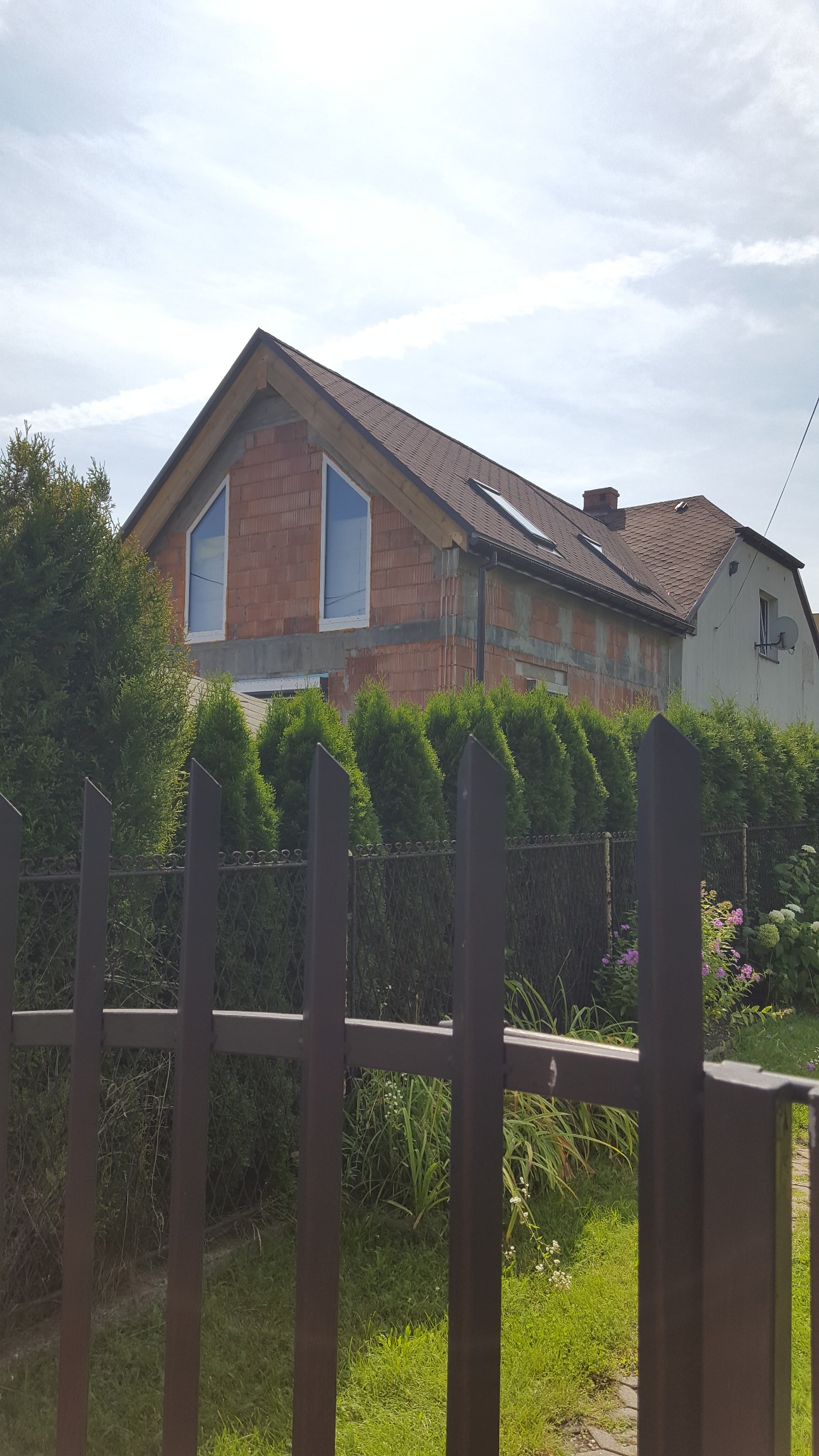 Dom w trakcie budowy z widocznymi bloczkami i oknami, częściowo zasłonięty przez żywopłot i ogrodzenie. W tle widoczny fragment drugiego, starszego domu.