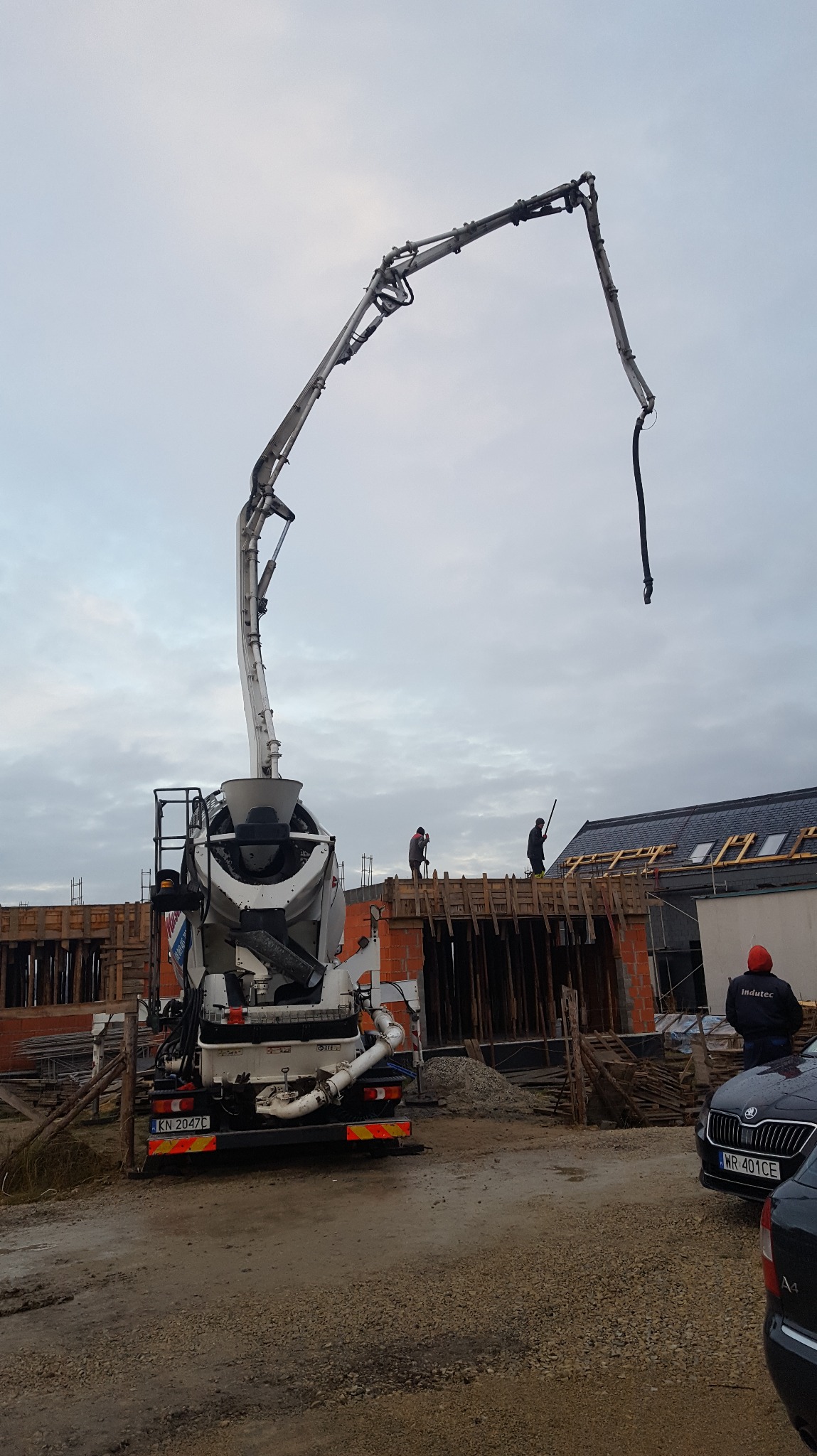 Betonomieszarka z pompą na budowie domu z cegły. Widoczni robotnicy w trakcie pracy, szalunki i zbrojenia. Pochmurne niebo w tle.