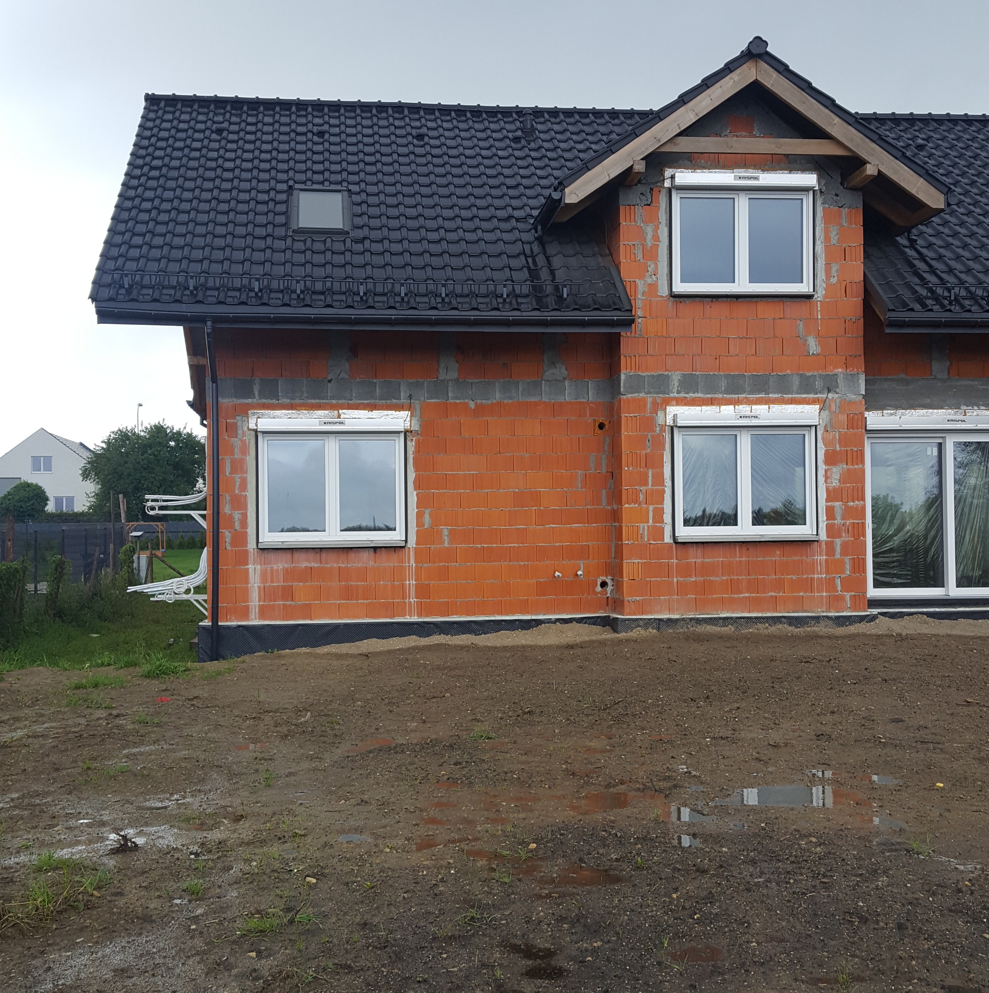 Niedokończony dom z czerwonej cegły z czarnym dachem, okna z roletami. Widoczne rury na zewnątrz budynku. Teren wokół domu jest błotnisty.