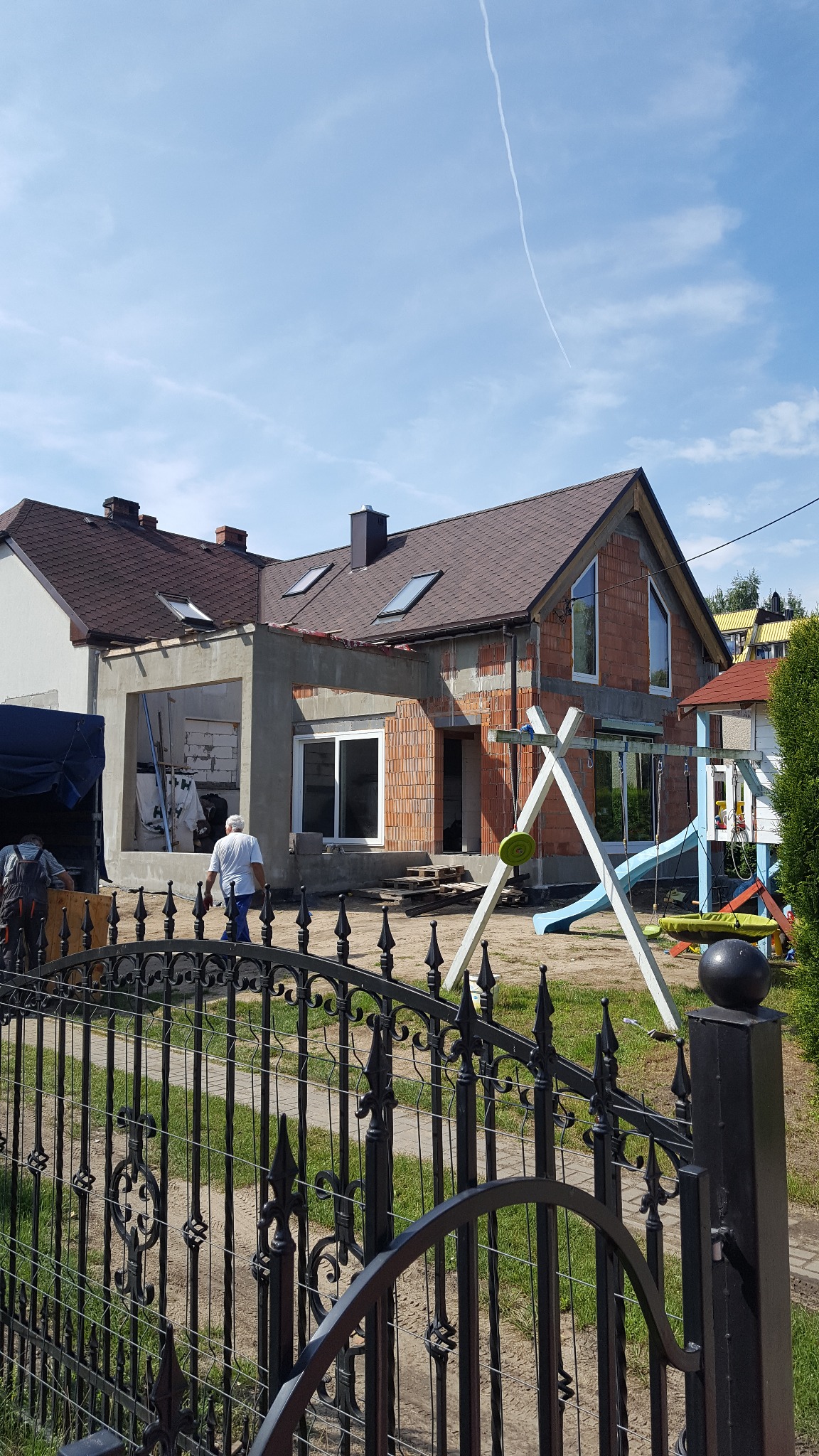 Rozbudowa domu z czerwonej cegły, widok zza ozdobnego, czarnego ogrodzenia. Dodana część w stanie surowym, widoczne okna i dach. Plac zabaw w tle.
