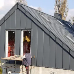 Montaż blachy na dachu skośnym z oknami dachowymi, widoczny pracownik na rusztowaniu i materiały izolacyjne wewnątrz budynku.