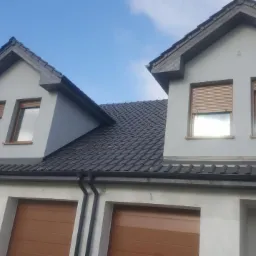 Nowy, ciemnoszary dach z dachówki ceramicznej na dwukondygnacyjnym domu z jasną elewacją, widoczne okna z roletami zewnętrznymi i fragment rusztowania.