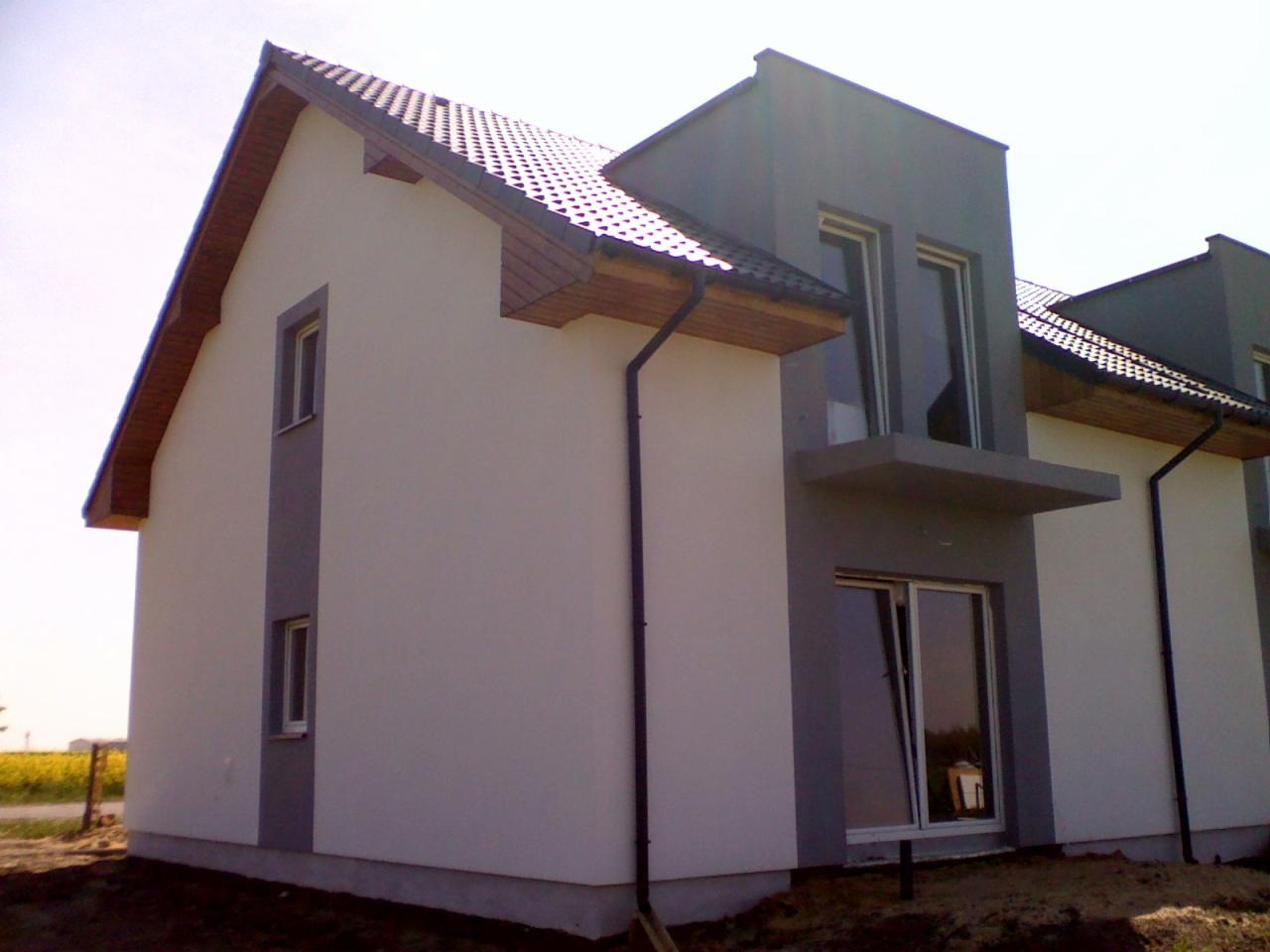 Nowoczesny dom jednorodzinny z dwuspadowym dachem pokrytym ciemną dachówką, z elementami elewacji w odcieniach szarości i bieli, widok z boku na tle pola.