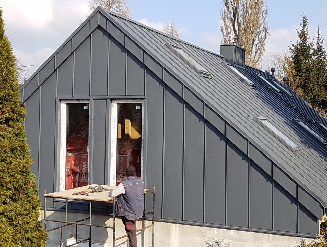 Montaż blachy na dachu skośnym z oknami dachowymi, widoczny pracownik na rusztowaniu i materiały izolacyjne wewnątrz budynku.