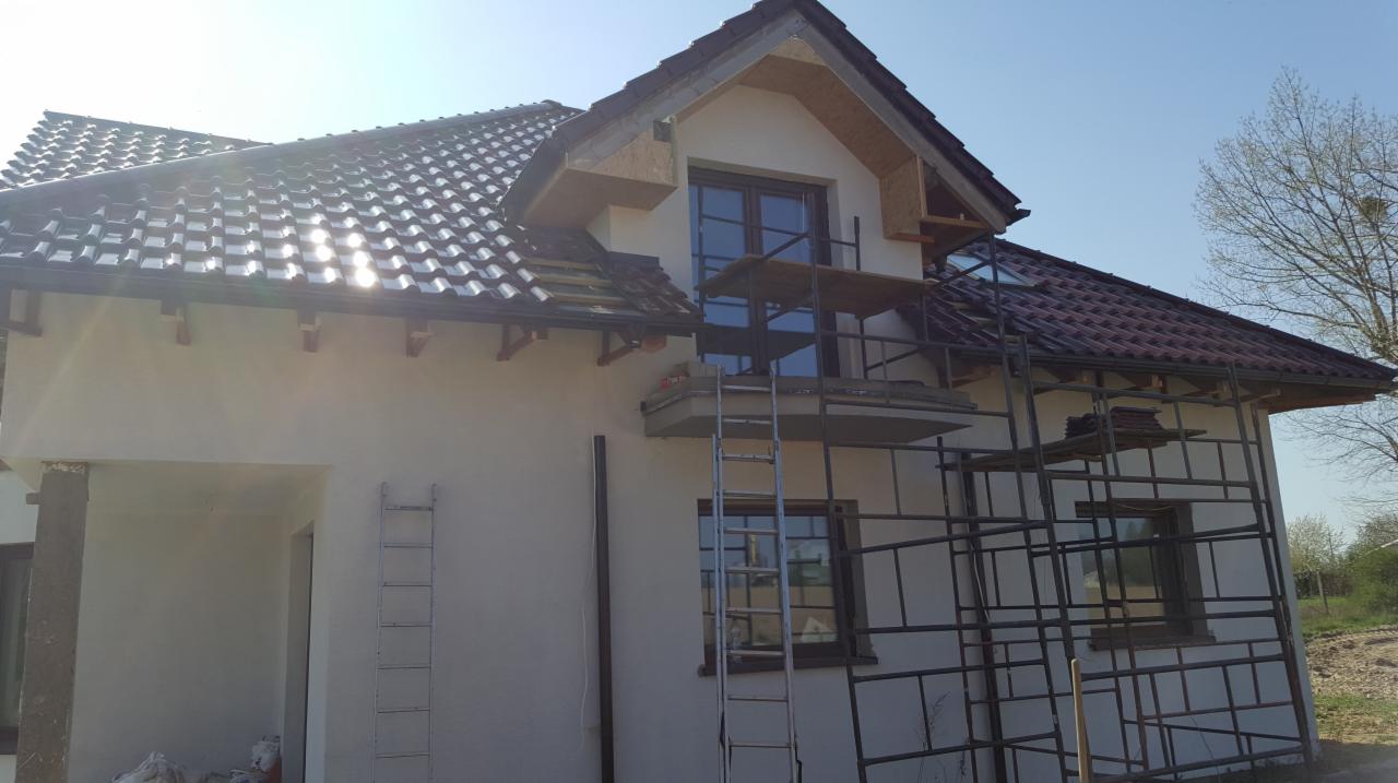 Dom w trakcie prac dekarskich, widoczne rusztowania, częściowo pokryty dach z ciemnej dachówki, przygotowane elementy konstrukcyjne nad oknem balkonowym.