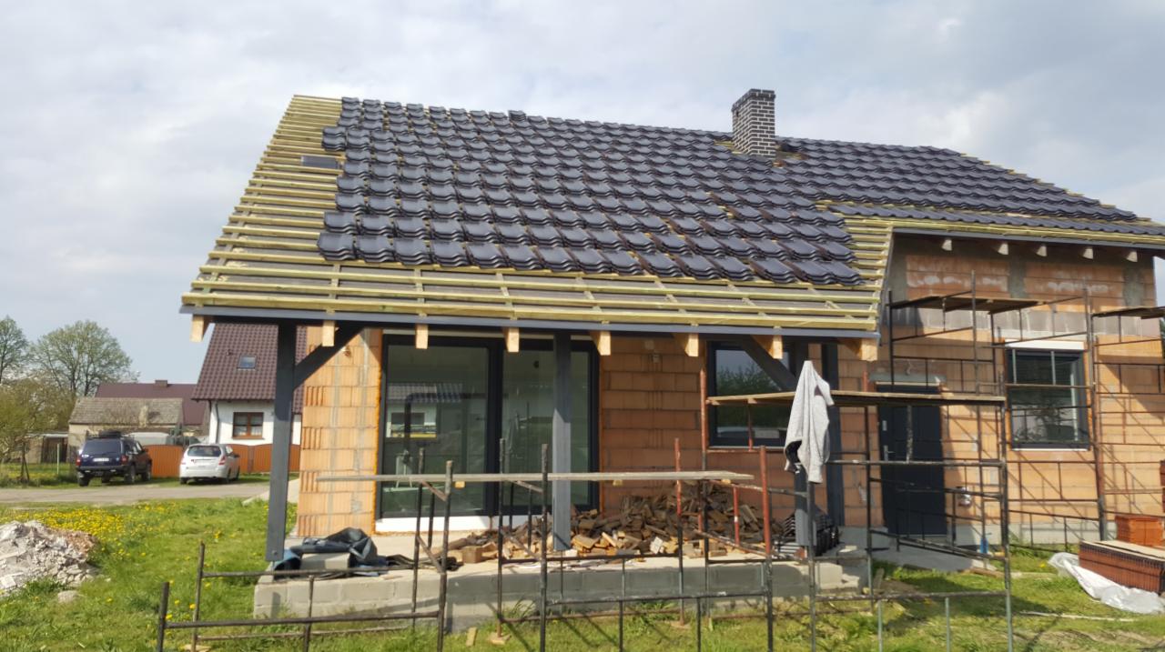 Montaż ciemnoszarej dachówki ceramicznej na nowym domu w trakcie budowy, widoczne rusztowania i stos drewna.