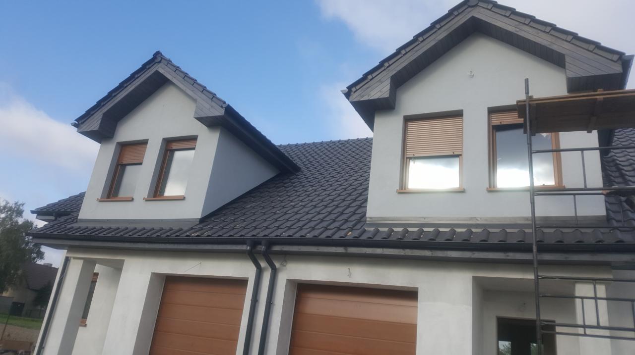 Nowy, ciemnoszary dach z dachówki ceramicznej na dwukondygnacyjnym domu z jasną elewacją, widoczne okna z roletami zewnętrznymi i fragment rusztowania.