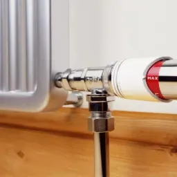 Chromowany zawór termostatyczny grzejnika z ustawieniem MAX, zamontowany na żeberkowym grzejniku panelowym, na tle drewnianej podłogi i białej ściany.