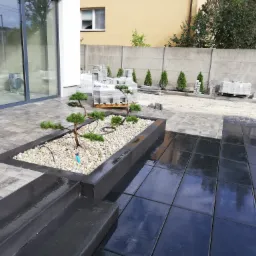 Nowoczesny taras z szarej kostki brukowej i czarnych płyt, z podwyższonym rabatem obsadzonym miniaturowymi drzewkami bonsai i białym grysem, w tle palety z kostką brukową i rząd małych tuj