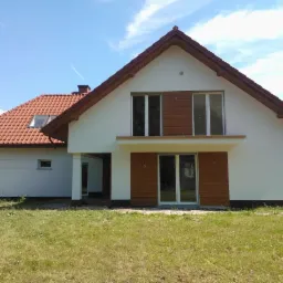 Nowoczesny dom z białą elewacją i drewnianymi panelami okiennymi, czerwony dach, zielony trawnik. Jasne, słoneczne ujęcie domu jednorodzinnego.