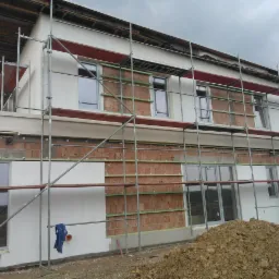 Dwupiętrowy budynek w trakcie prac elewacyjnych, widoczne rusztowania, częściowo otynkowana i ocieplona ściana z fragmentami odsłoniętej cegły, składowisko ziemi przed budynkiem.