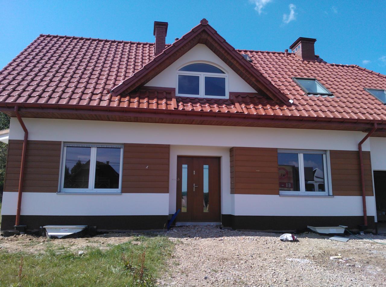 Dom jednorodzinny z elewacją w kolorze białym i brązowym, z czerwonym dachem i oknami. Widoczne rynny i fragment terenu przed budynkiem.