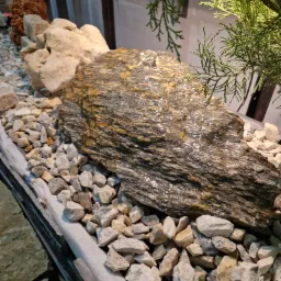 Dekoracyjny ogródek skalny z dużym, błyszczącym kamieniem o warstwowej strukturze, otoczonym drobnymi, białymi kamykami i gałązkami iglastego krzewu.