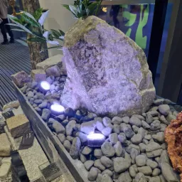 Dekoracyjna kompozycja z dużego, podświetlonego kamienia, otoczona drobnymi otoczakami i roślinnością, z widocznymi elementami ekspozycji budowlanej w tle.