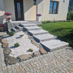 Schody wejściowe do domu wyłożone szarą kostką granitową, z białym żwirem i ozdobnymi kamieniami oraz roślinami, prowadzące od chodnika z kostki do drzwi wejściowych.