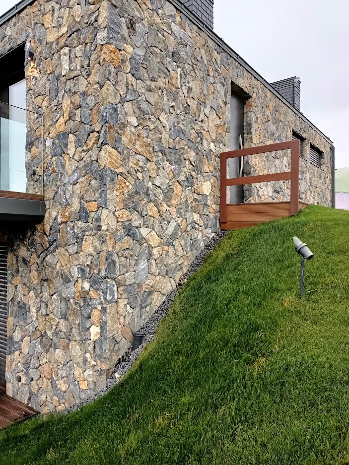 Elewacja domu wykończona nieregularnym kamieniem w odcieniach szarości i beżu, z trawiastym wzgórzem i oświetleniem zewnętrznym. Widoczny fragment tarasu z drewnianą balustradą.
