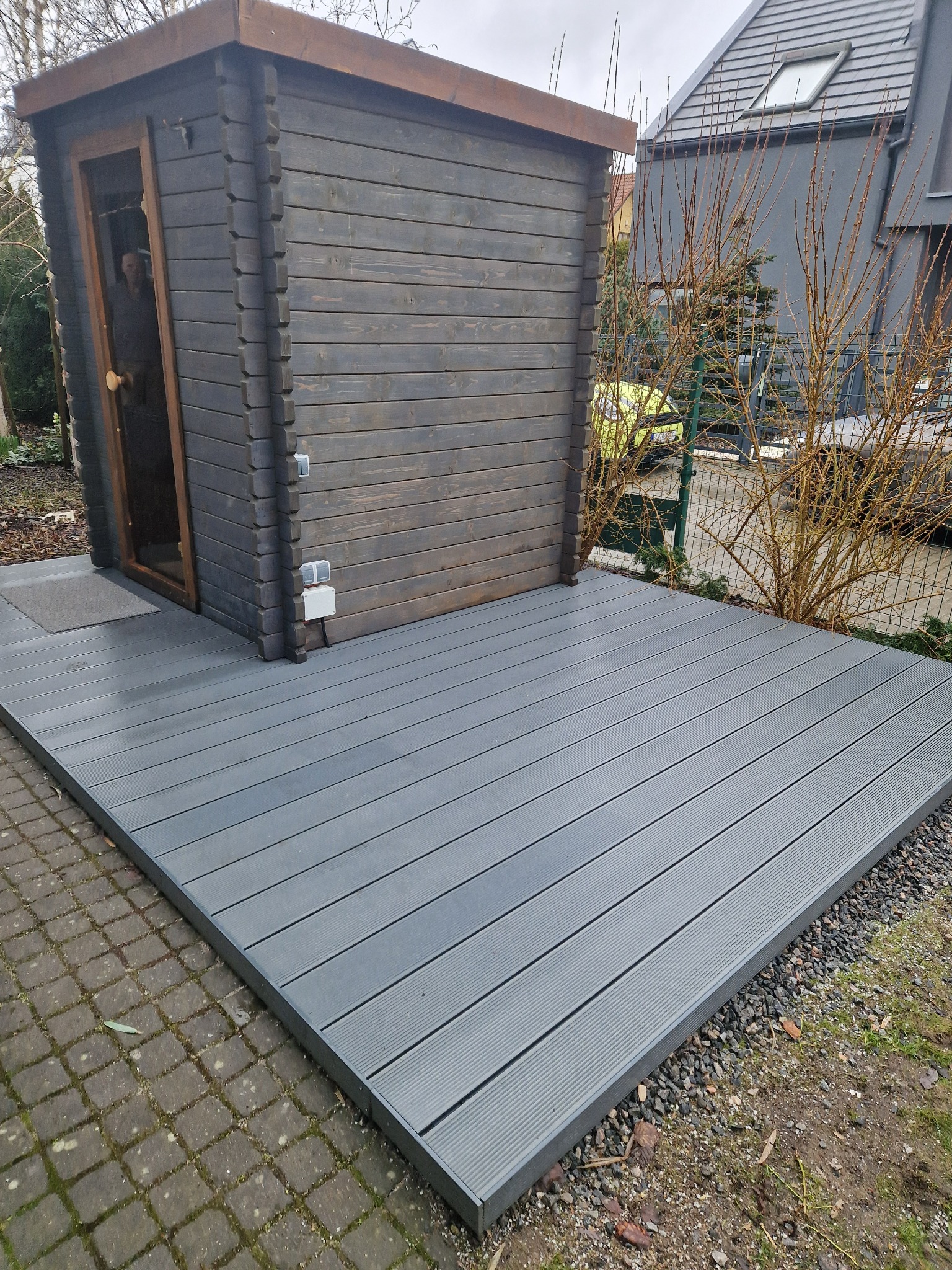 Szara deska kompozytowa tarasu przylegająca do ciemnej drewnianej altany z widoczną osobą w środku, obok kostka brukowa i krzewy bez liści.