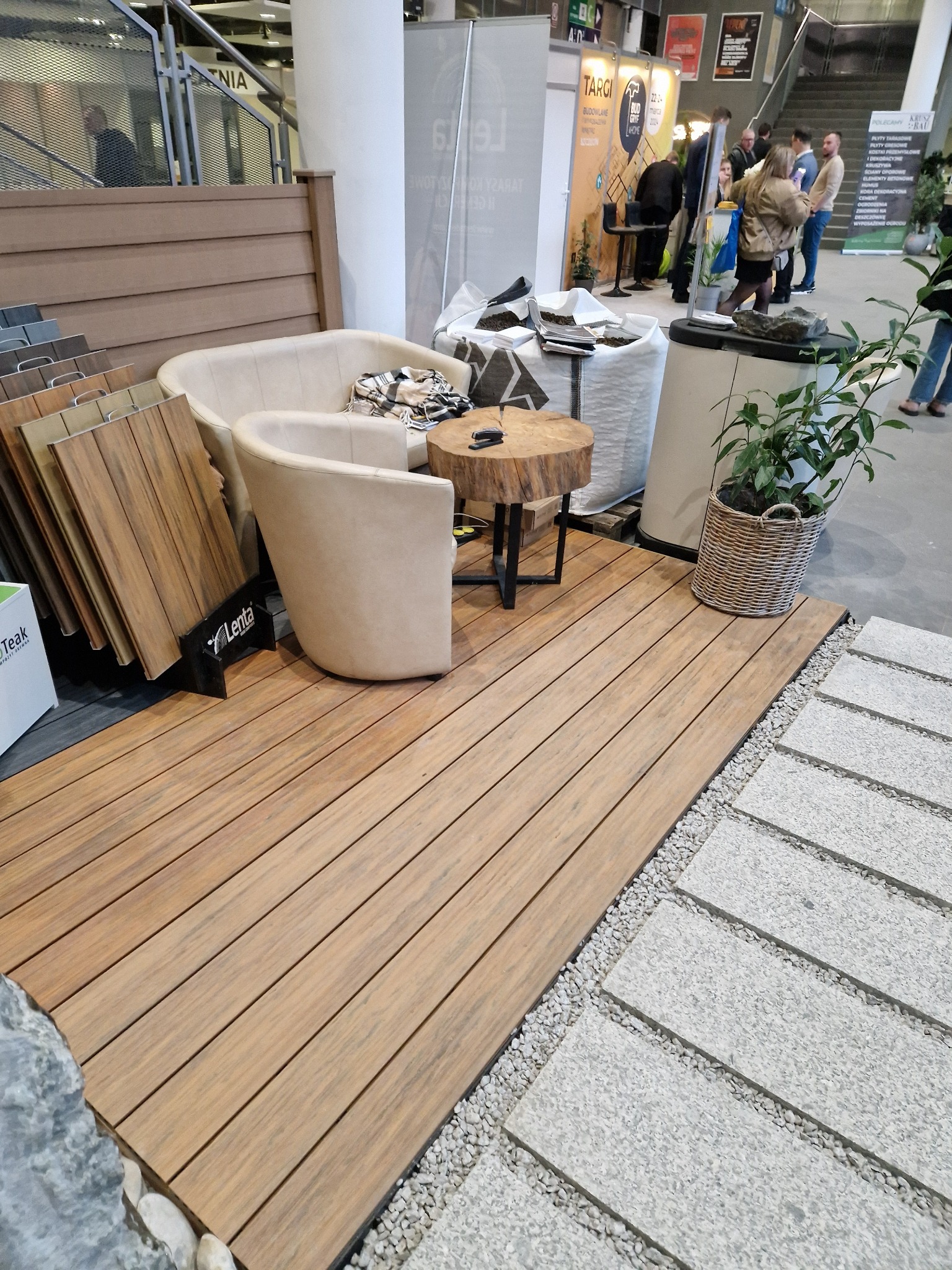 Aranżacja tarasu kompozytowego z meblami ogrodowymi na tle ekspozycji targowej, widoczne próbki materiałów i rośliny w koszu.