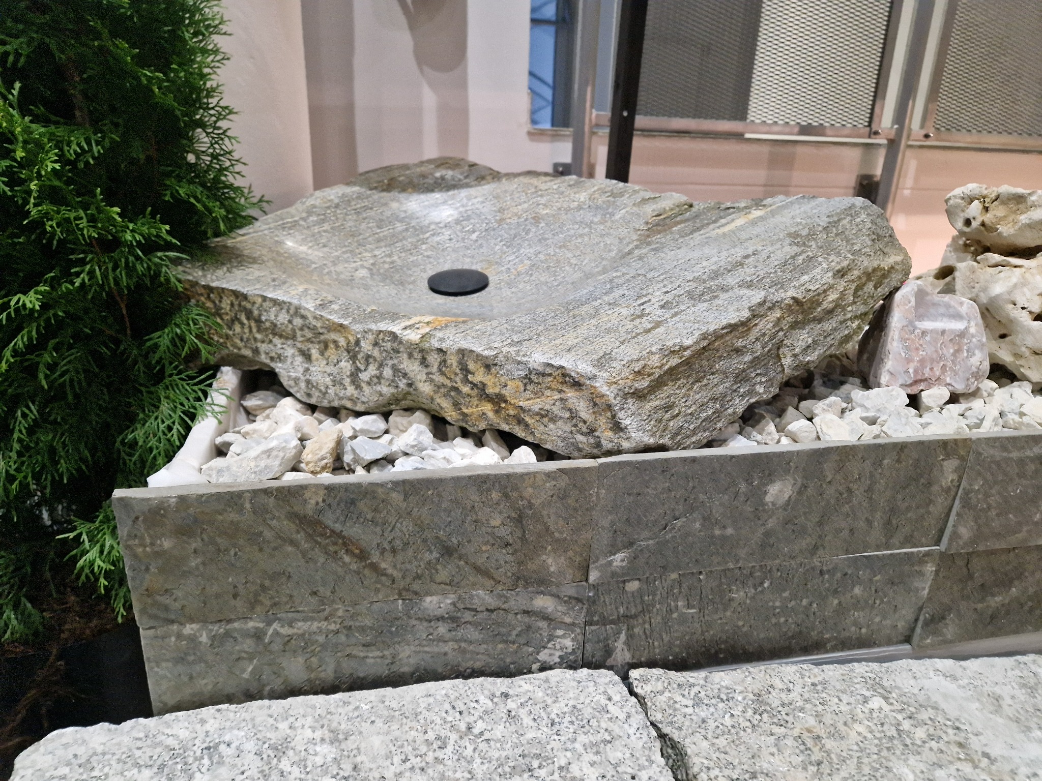 Kamienna umywalka z odpływem, otoczona białymi kamykami i zieloną rośliną, na tle ściany z metalową kratą.