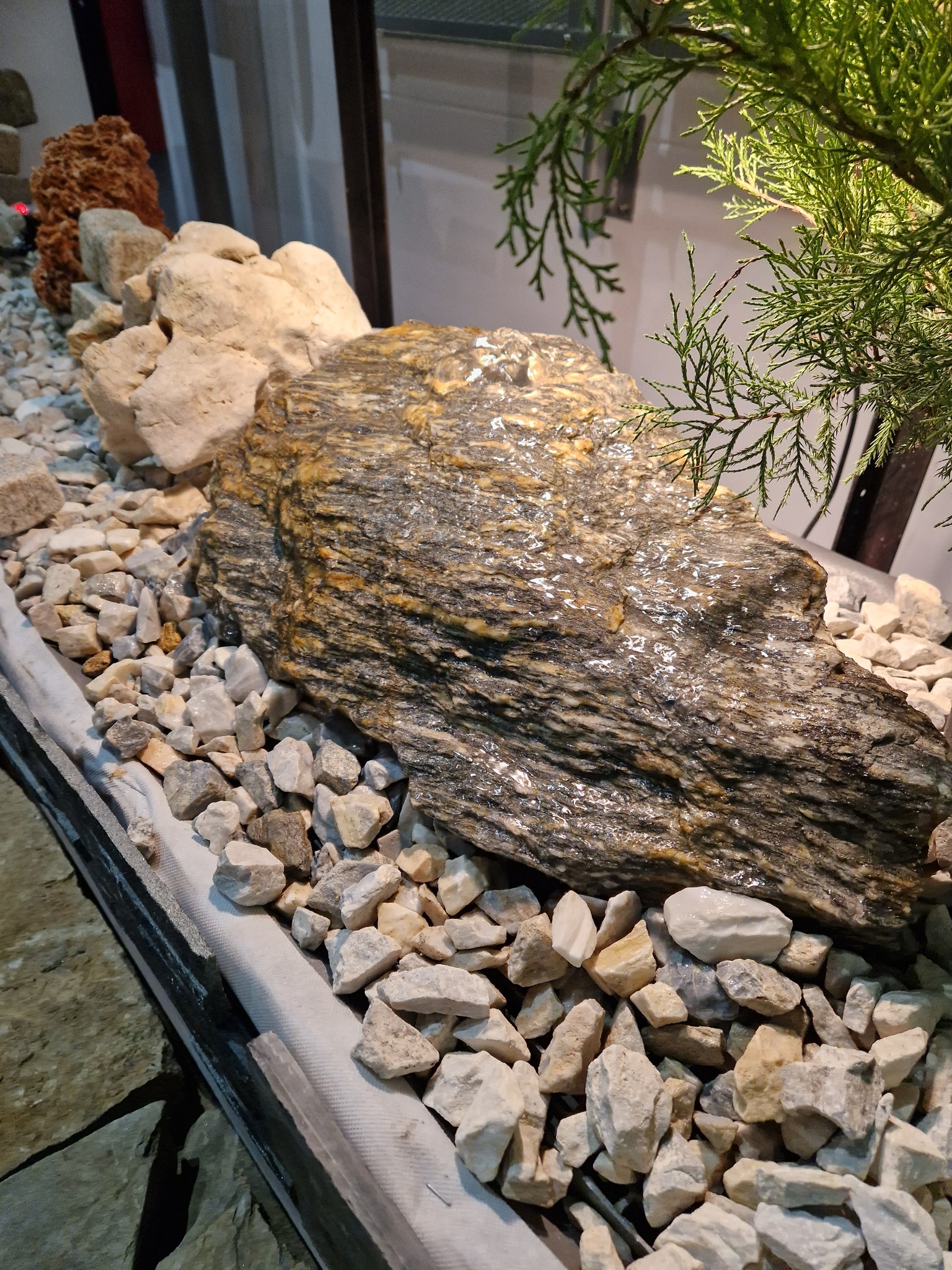 Dekoracyjny ogródek skalny z dużym, błyszczącym kamieniem o warstwowej strukturze, otoczonym drobnymi, białymi kamykami i gałązkami iglastego krzewu.