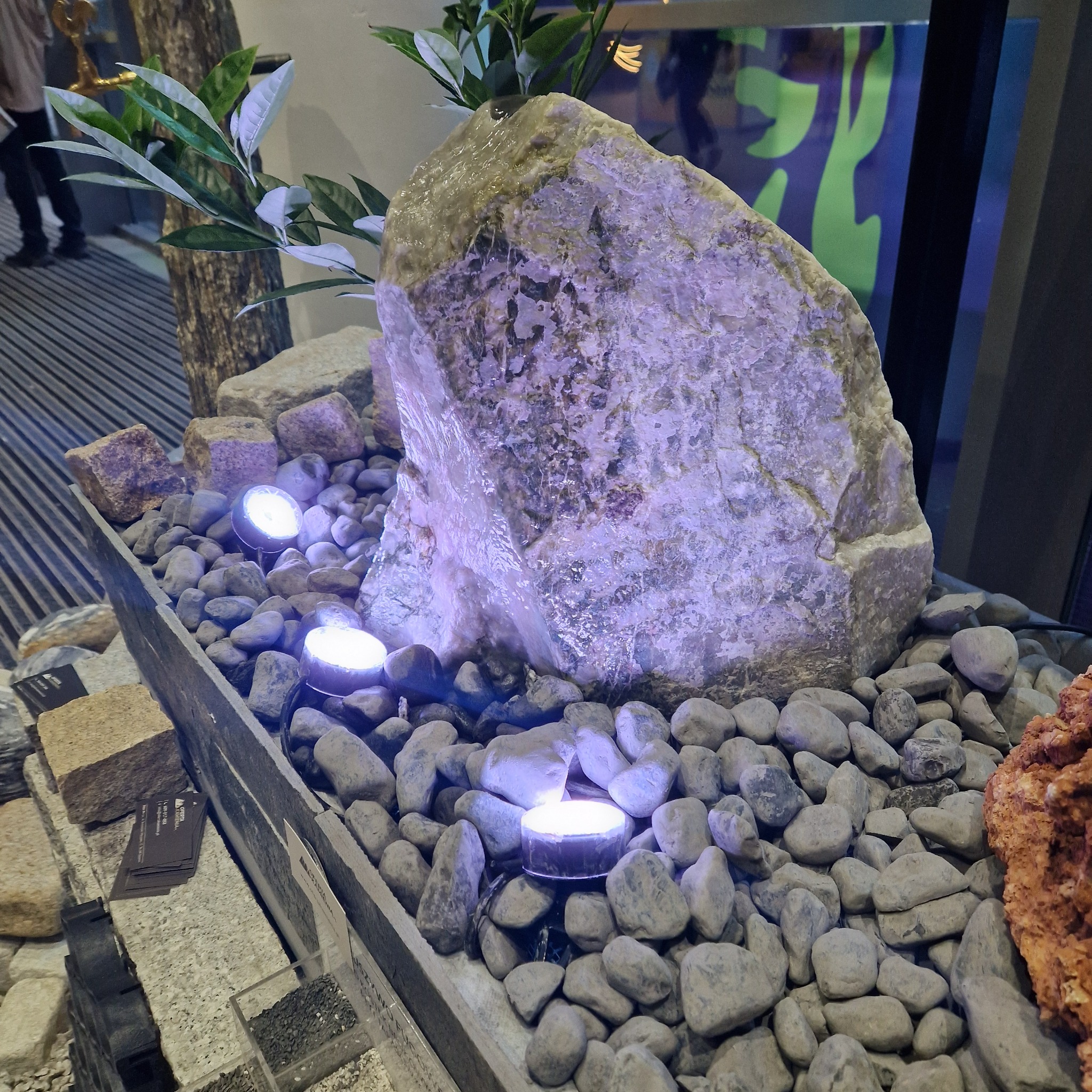 Dekoracyjna kompozycja z dużego, podświetlonego kamienia, otoczona drobnymi otoczakami i roślinnością, z widocznymi elementami ekspozycji budowlanej w tle.