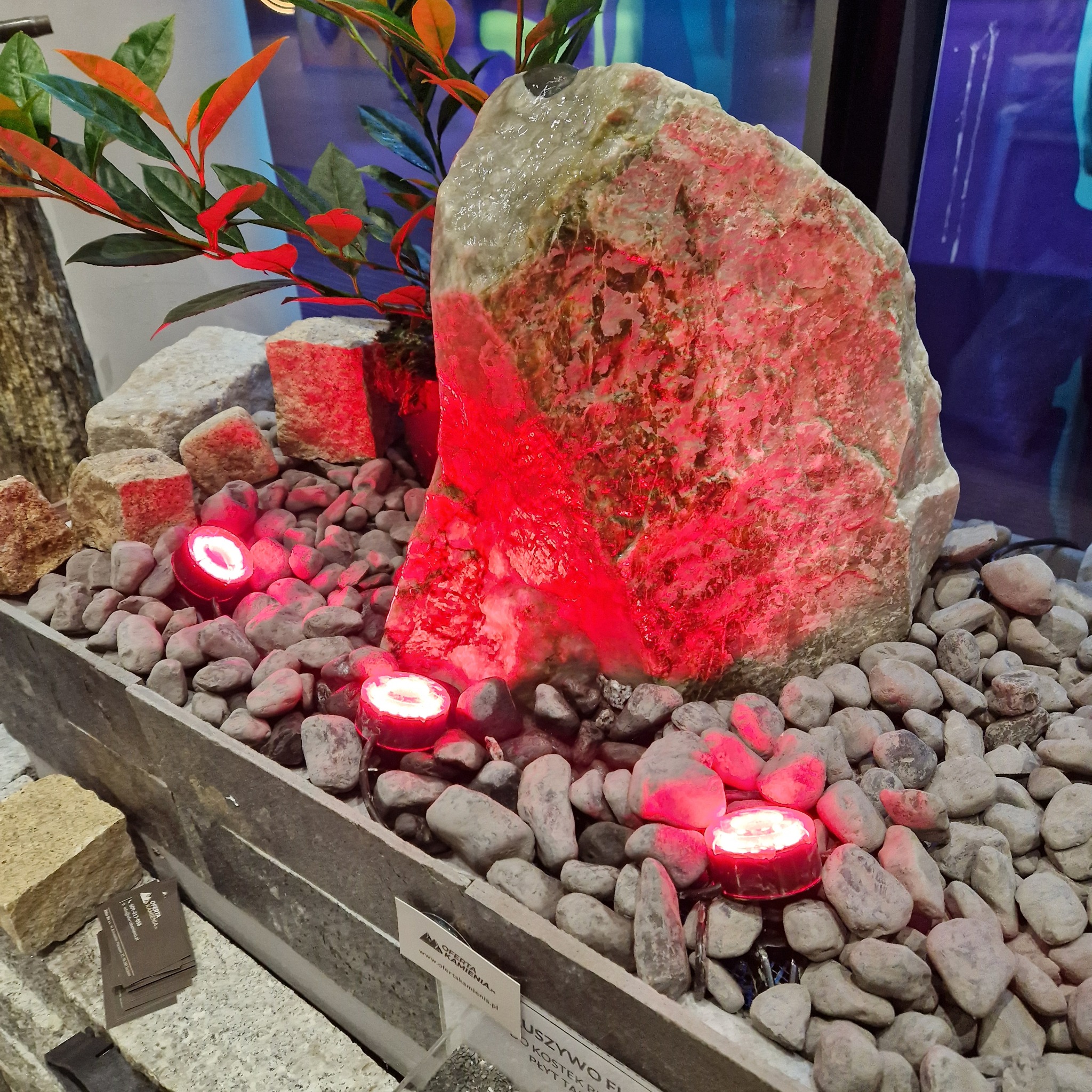 Dekoracyjna kompozycja z dużego kamienia podświetlonego na czerwono, otoczonego drobnymi otoczakami i sztuczną roślinnością, prezentowana w szarej, kamiennej donicy z oświetleniem LED.