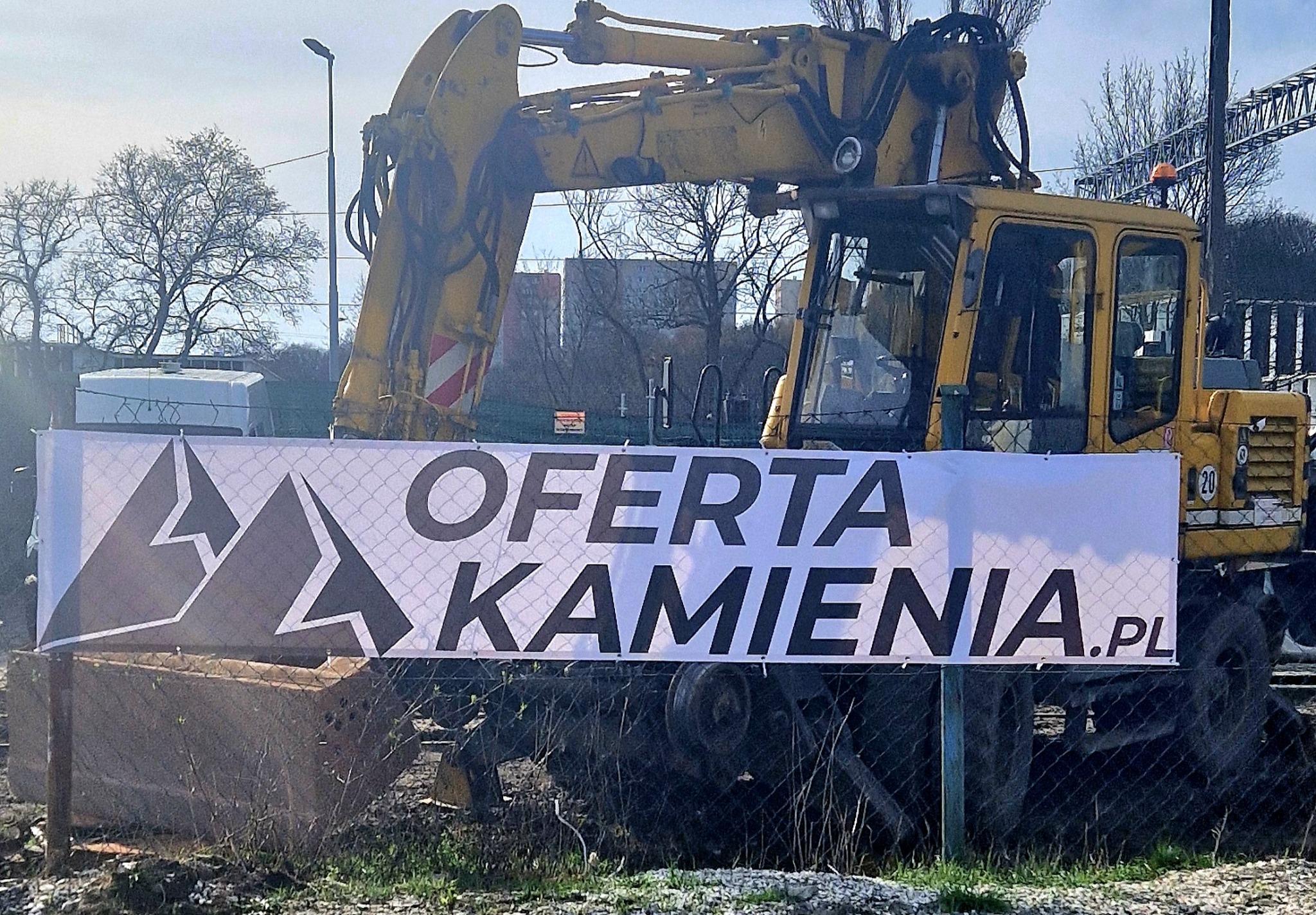 Żółta koparka z banerem reklamowym 'Oferta Kamienia.pl' na tle placu budowy z widocznymi drzewami i budynkami w oddali.