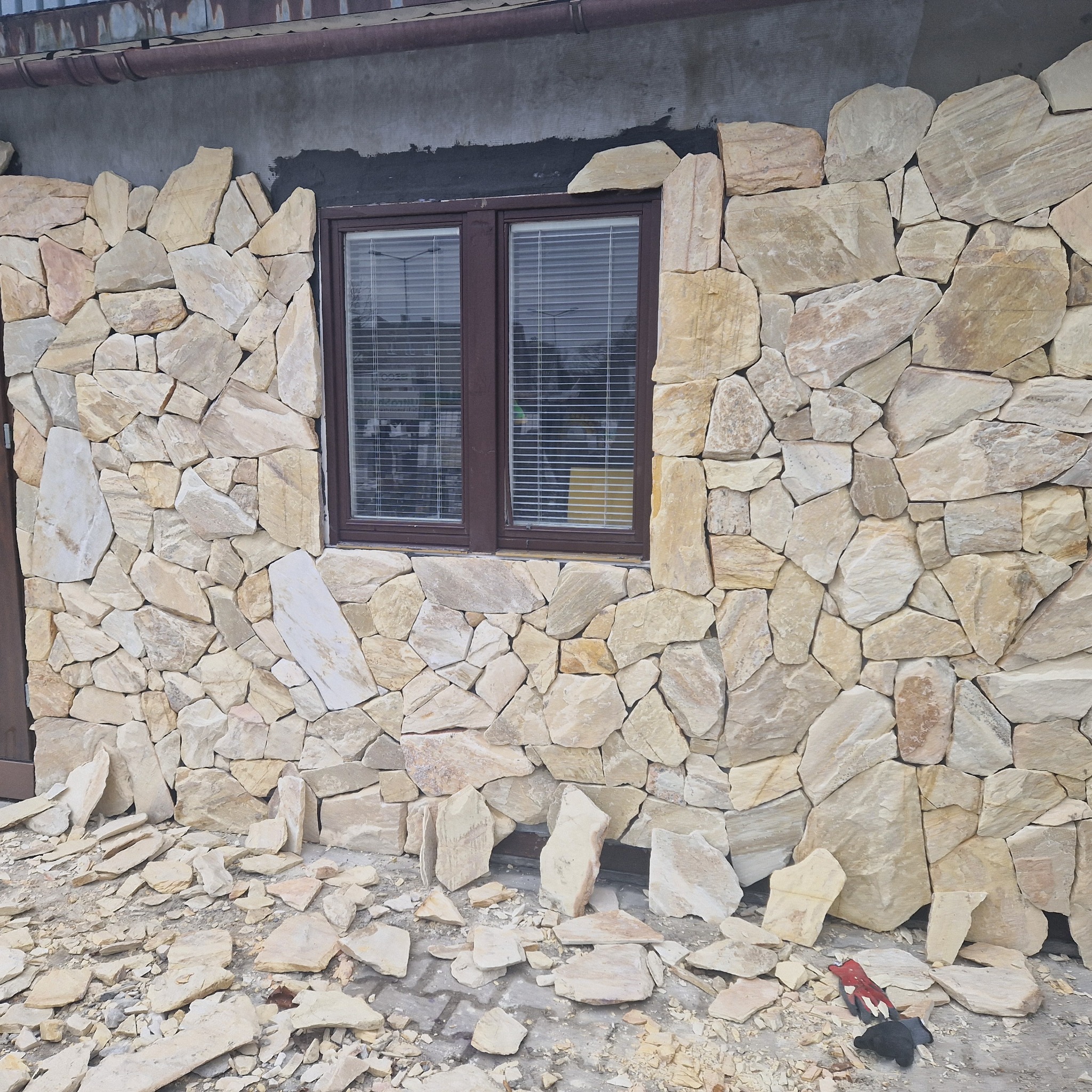 Montaż elewacji z nieregularnych kamieni piaskowca wokół okna z żaluzjami, widoczne fragmenty zaprawy i odłamki kamienia na ziemi.