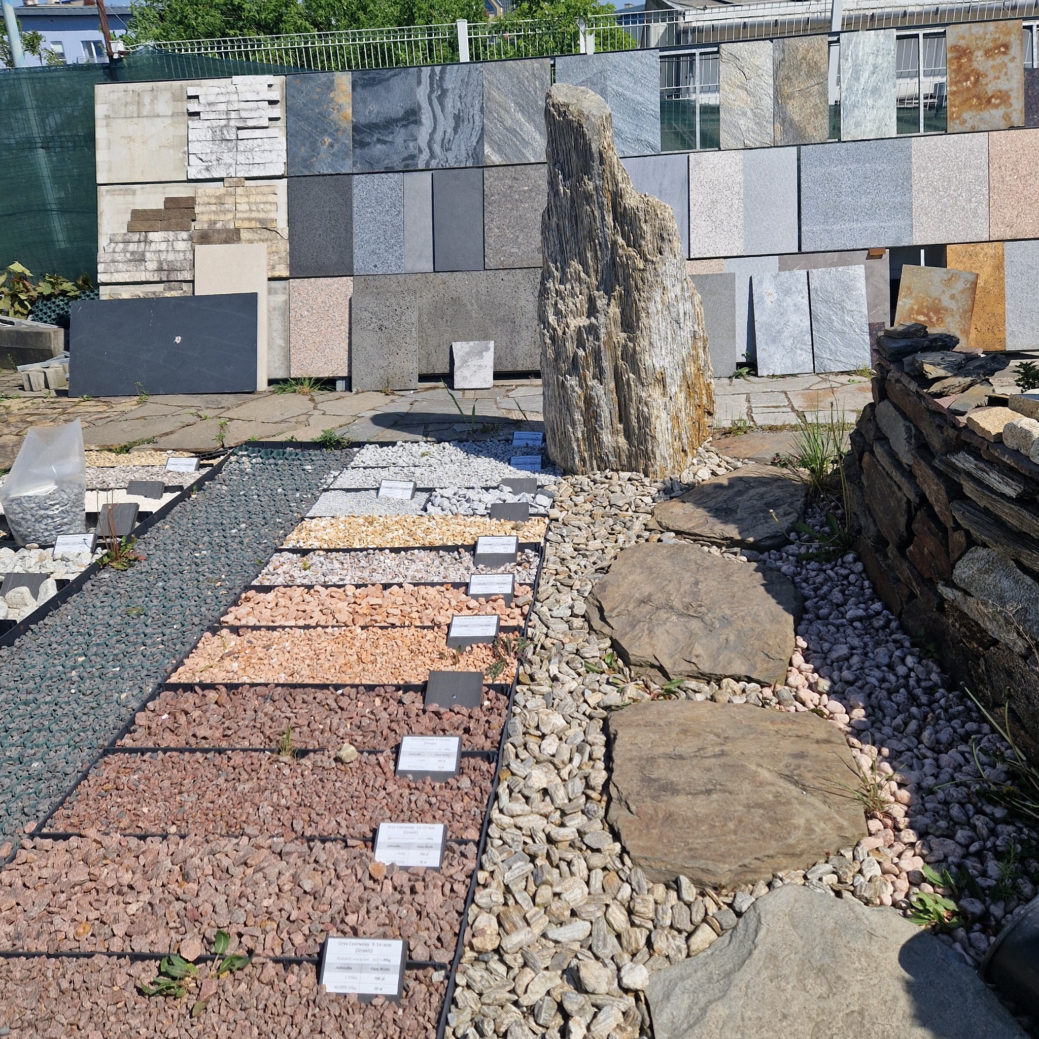 Ekspozycja różnorodnych kruszyw i płyt kamiennych na zewnątrz, z pionową skałą dekoracyjną i fragmentem murku z łupka w tle.