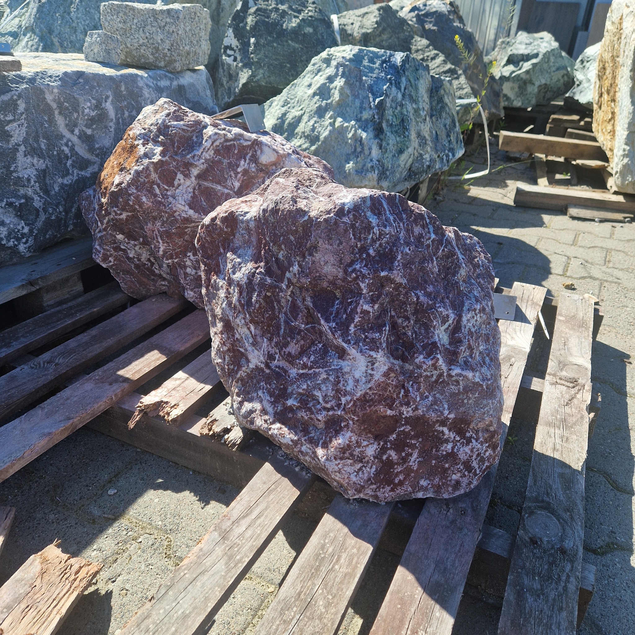 Dwa duże, czerwono-białe kamienie dekoracyjne na drewnianej palecie w Szczecinie, otoczone innymi, szarymi kamieniami o różnej wielkości.