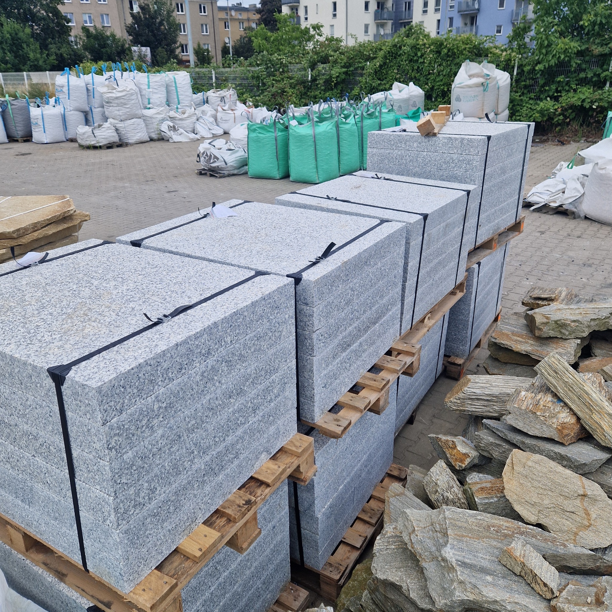 Palety z szarą kostką brukową na placu budowy, obok stosy kamieni łupanych i worki z materiałami budowlanymi w tle.