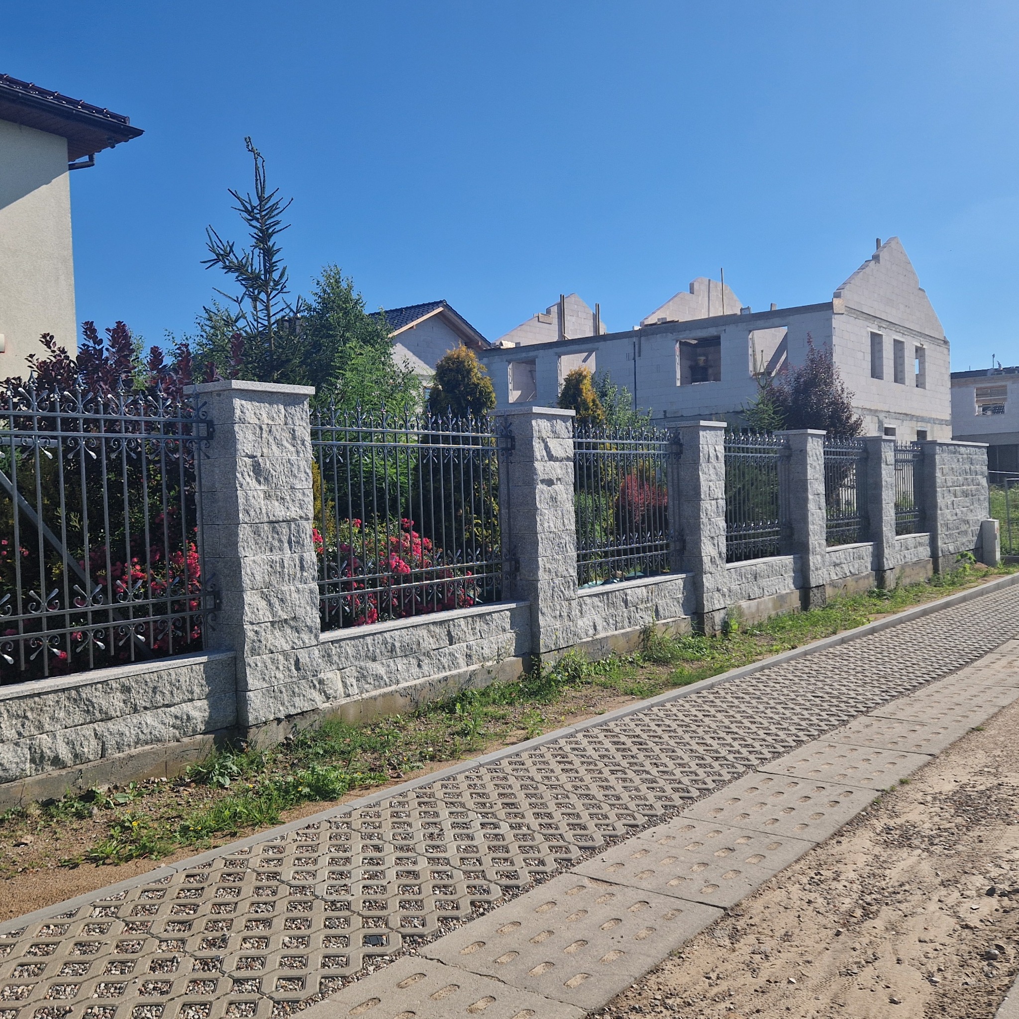 Ogrodzenie z szarej kostki z metalowymi przęsłami, oddzielające posesję z domem w budowie od chodnika z ażurowych płyt betonowych.
