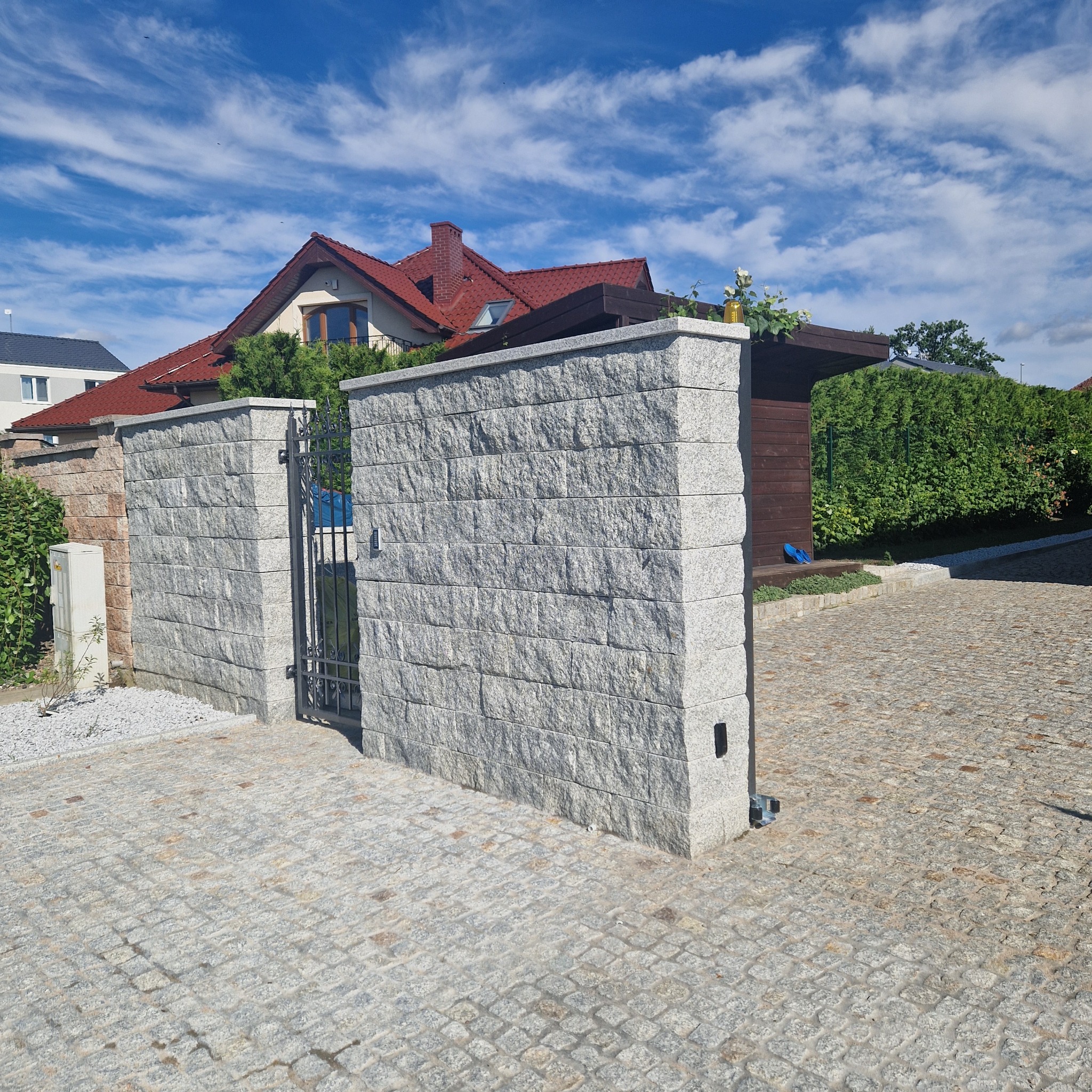 Ogrodzenie z szarej kostki, brama z kutego żelaza i nawierzchnia z kostki, widok na dom z czerwonym dachem w tle.