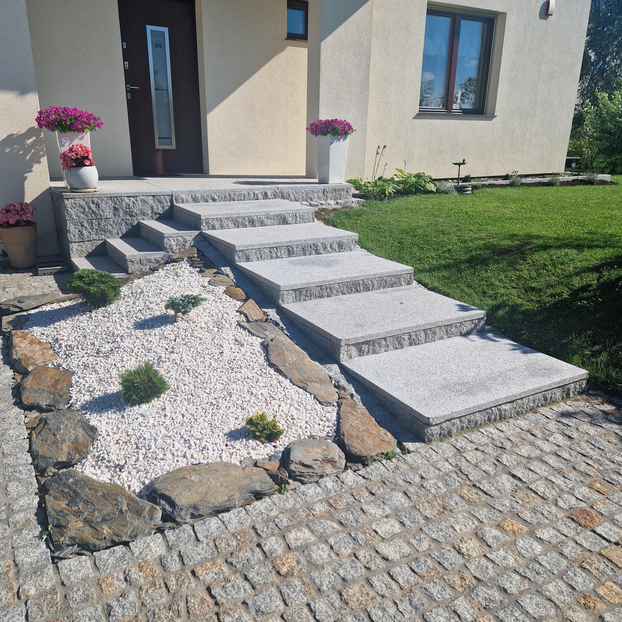 Schody wejściowe do domu wyłożone szarą kostką granitową, z białym żwirem i ozdobnymi kamieniami oraz roślinami, prowadzące od chodnika z kostki do drzwi wejściowych.