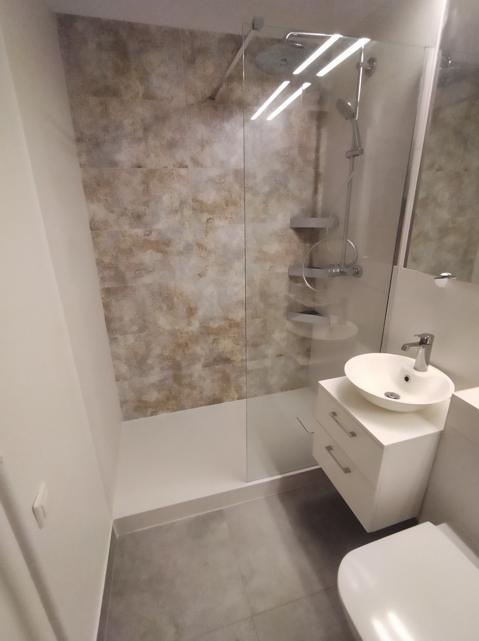 Nowoczesna łazienka po remoncie: kabina prysznicowa ze szklaną ścianką, umywalka nablatowa na szafce z szufladami, szare płytki podłogowe i beżowe płytki ścienne.