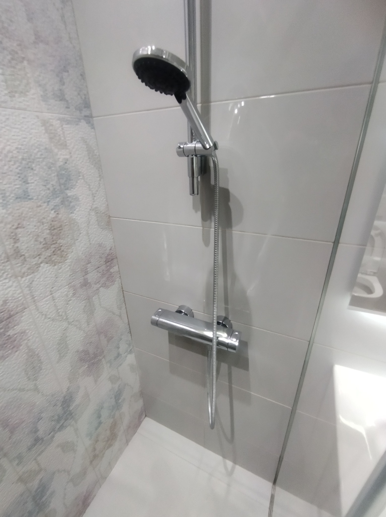 Nowoczesny zestaw prysznicowy z termostatem na tle białych i wzorzystych płytek w łazience. Chromowana armatura, minimalistyczny design.