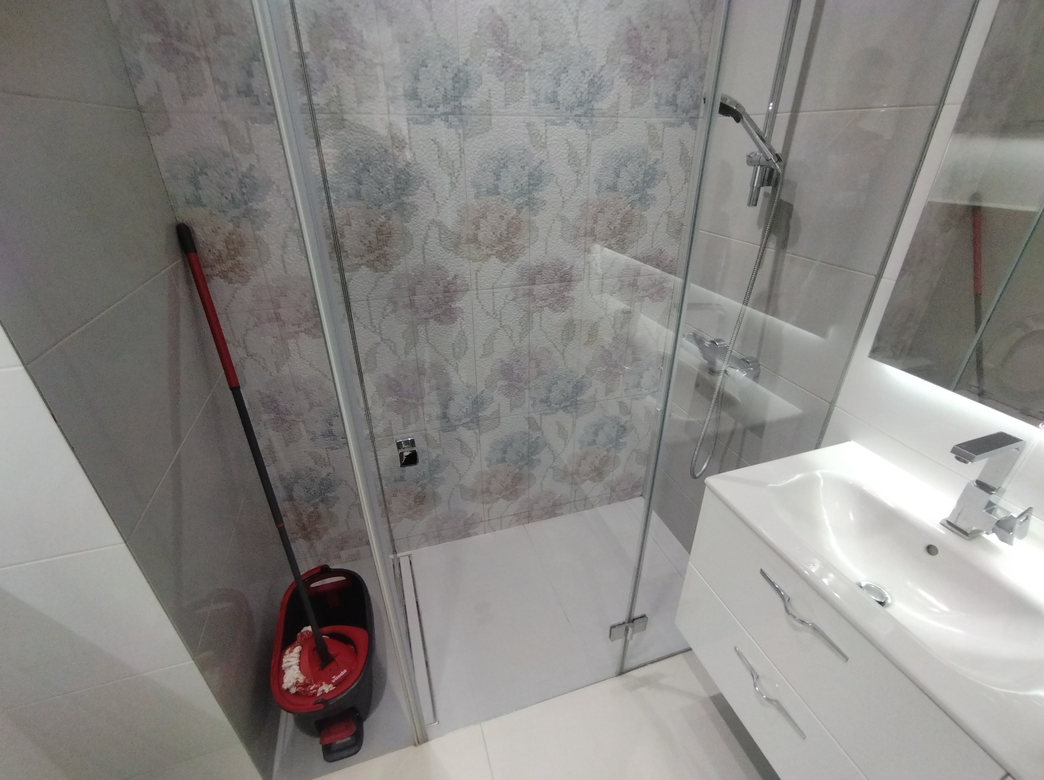 Nowoczesna kabina prysznicowa z kwiatowym motywem na płytkach, biała umywalka z szafką i mop w rogu. Minimalistyczny design łazienki po remoncie.