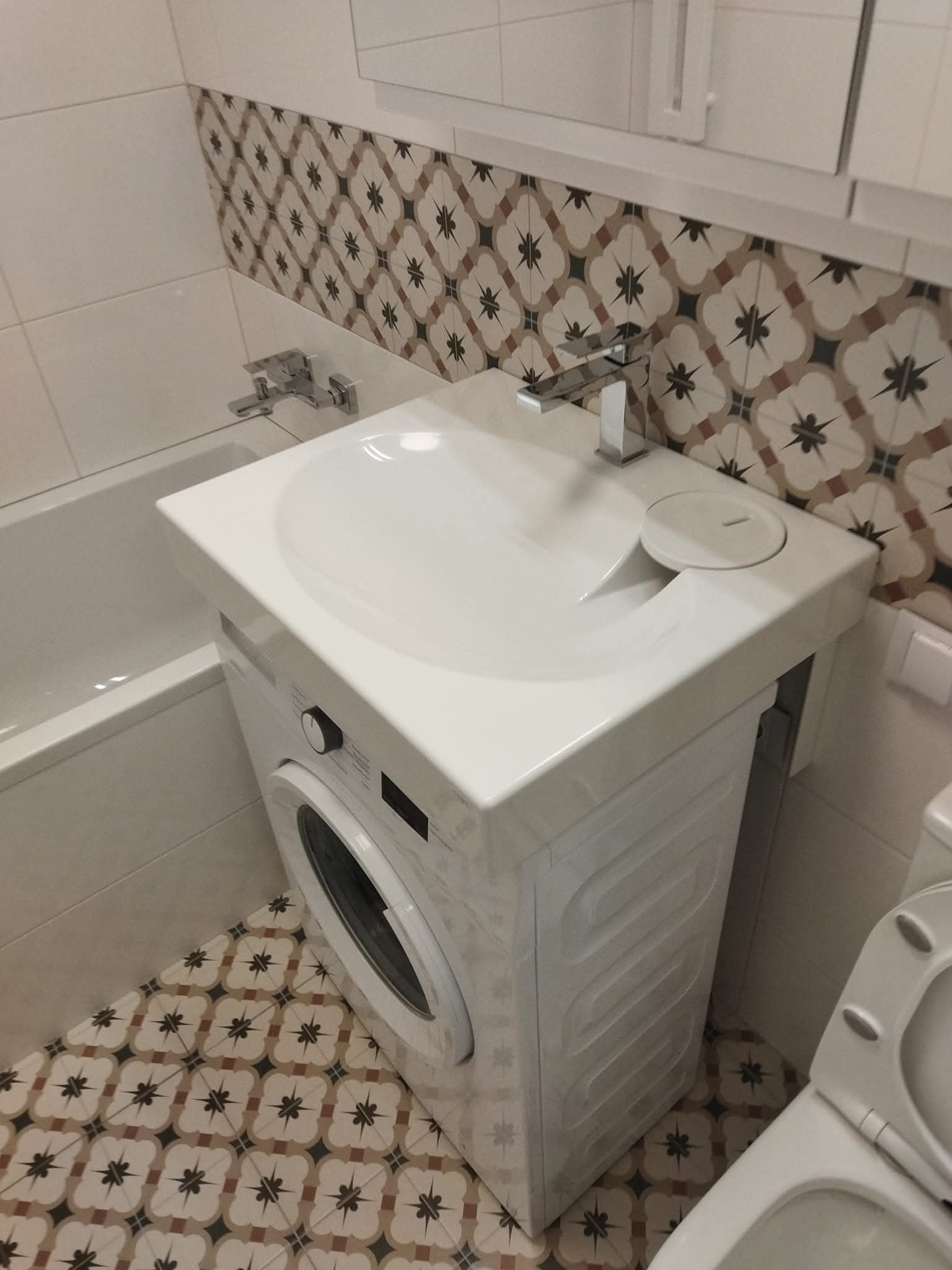 Nowoczesna łazienka: biała umywalka nad pralką, bateria chromowana, wzorzyste płytki na ścianie i podłodze. Kompaktowe rozwiązanie do małej przestrzeni.