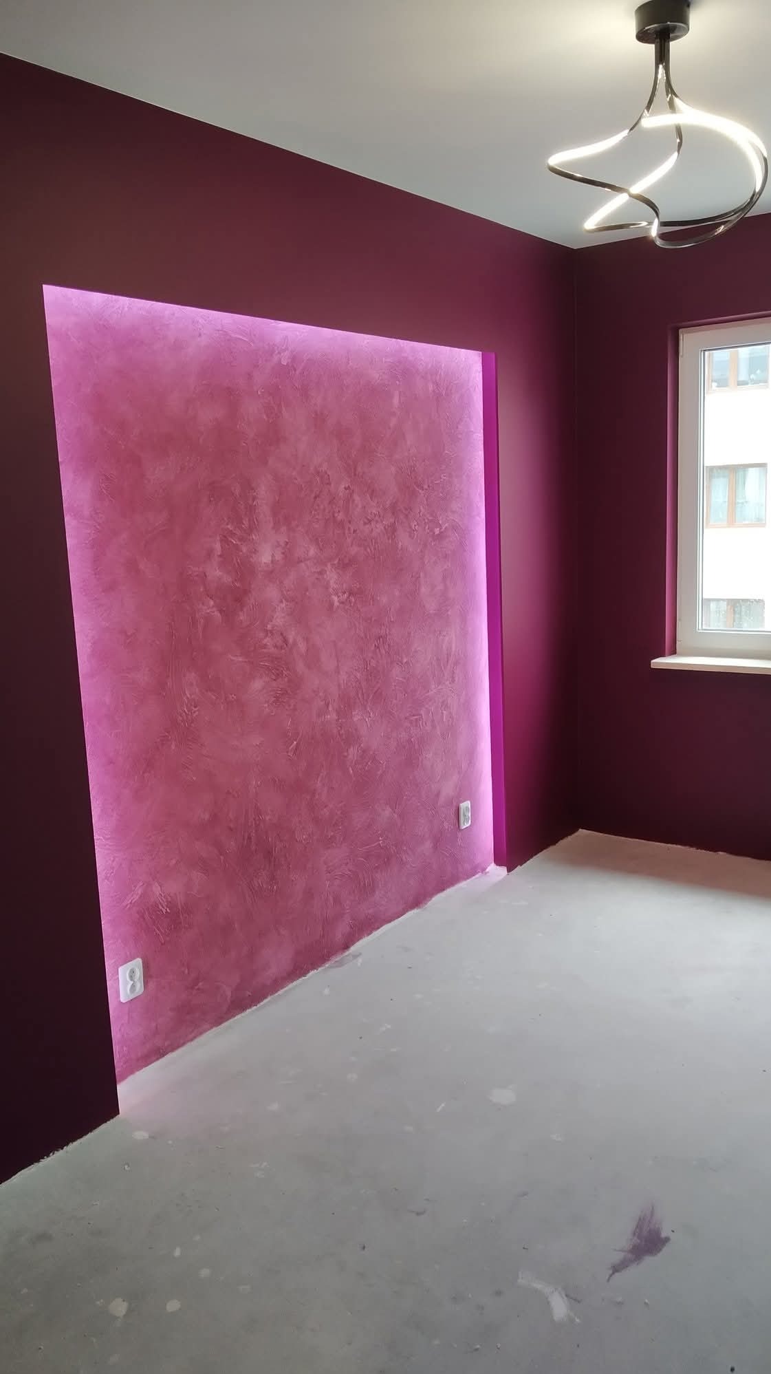 Fioletowa ściana z dekoracyjnym tynkiem i podświetleniem LED w nowocześnie wykończonym wnętrzu. Widoczny fragment okna i designerska lampa sufitowa.