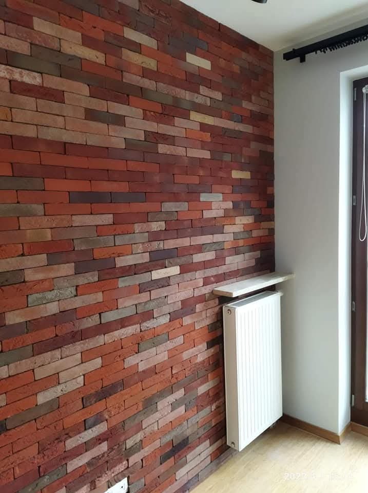 Ściana z cegły klinkierowej w odcieniach czerwieni i brązu, z grzejnikiem i parapetem. Widok z boku, ukazujący strukturę i kolorystykę cegieł.