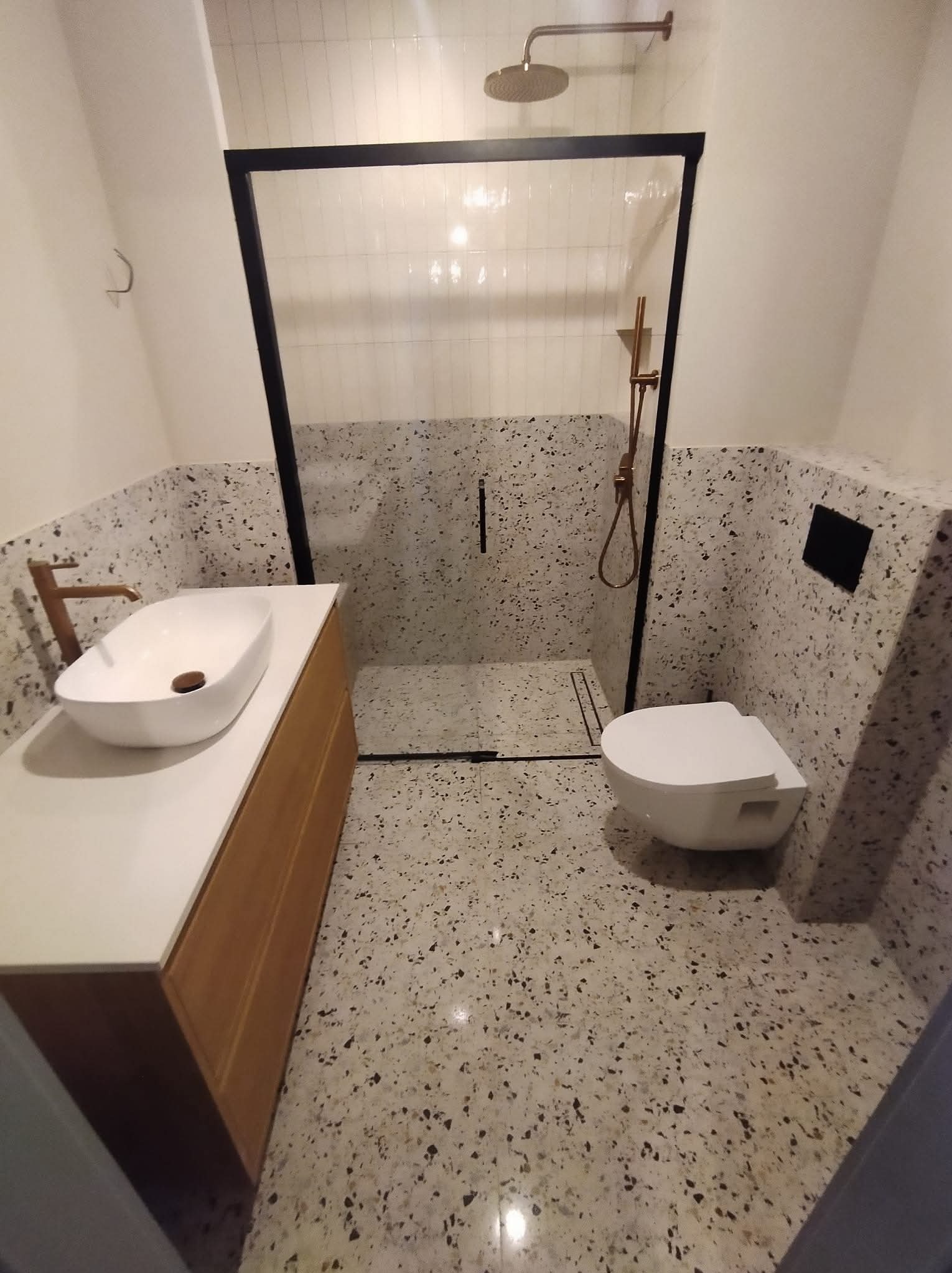 Nowoczesna łazienka z kabiną typu walk-in, umywalką nablatową i toaletą. Ściany i podłoga wykończone lastryko. Armatura w kolorze miedzi. Minimalistyczny design.