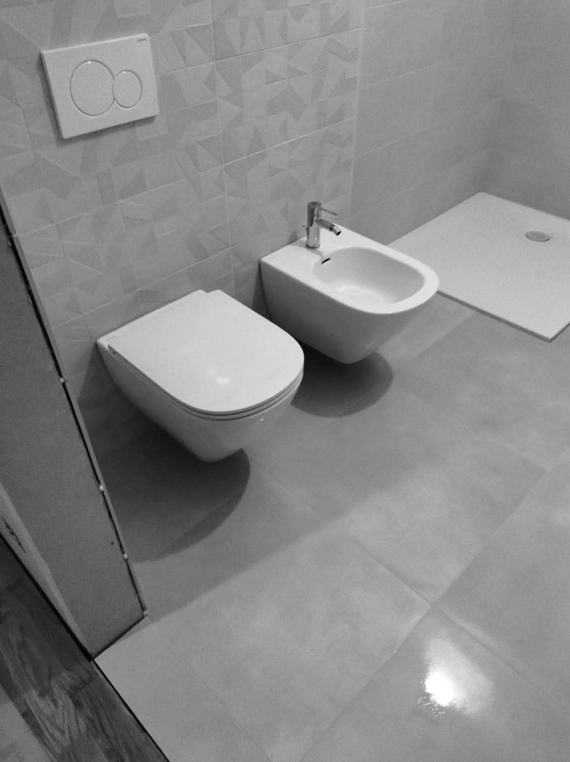 Nowoczesna, minimalistyczna łazienka w odcieniach szarości. Wisząca toaleta, bidet z baterią i brodzik tworzą spójną całość. Geometryczne płytki na ścianie dodają charakteru.