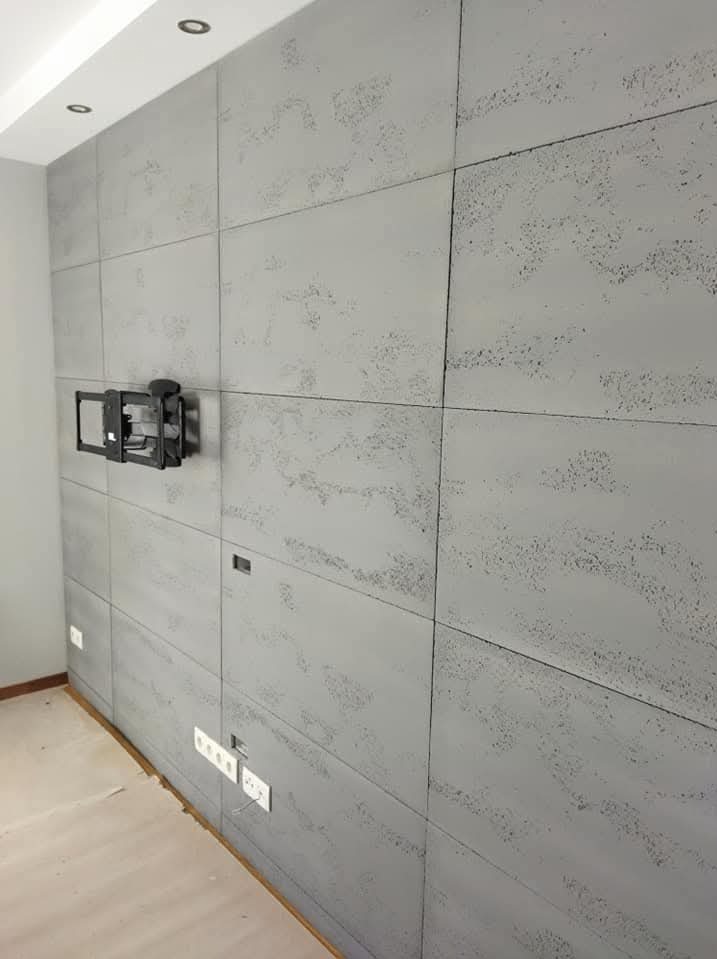 Ściana wykończona szarymi panelami imitującymi beton, z zamontowanym uchwytem na telewizor i gniazdkami elektrycznymi. Minimalistyczny design wnętrza.