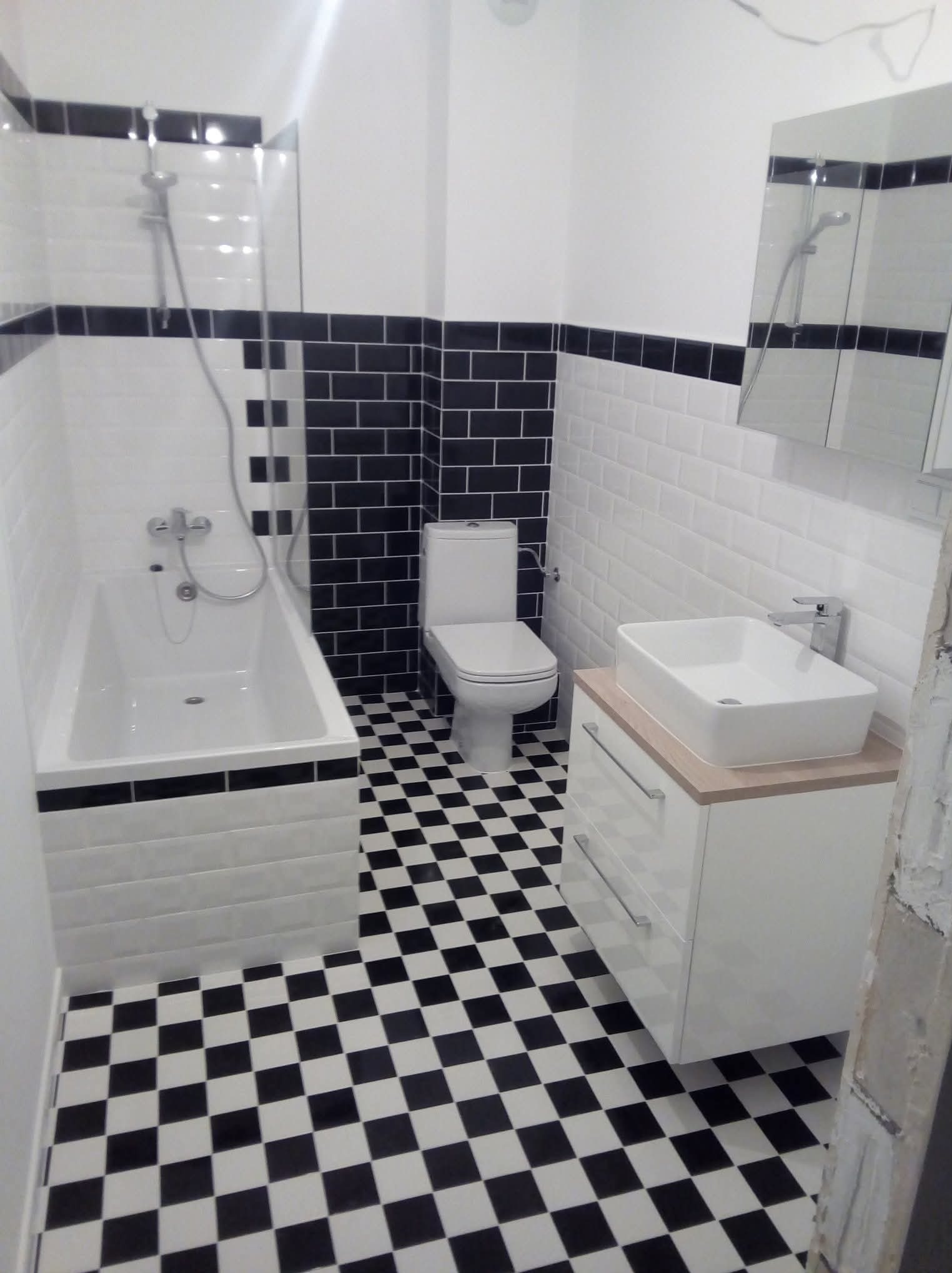 Wyremontowana łazienka w stylu retro: czarno-biała szachownica na podłodze, białe płytki z czarnym pasem, wanna, toaleta i umywalka z szafką.
