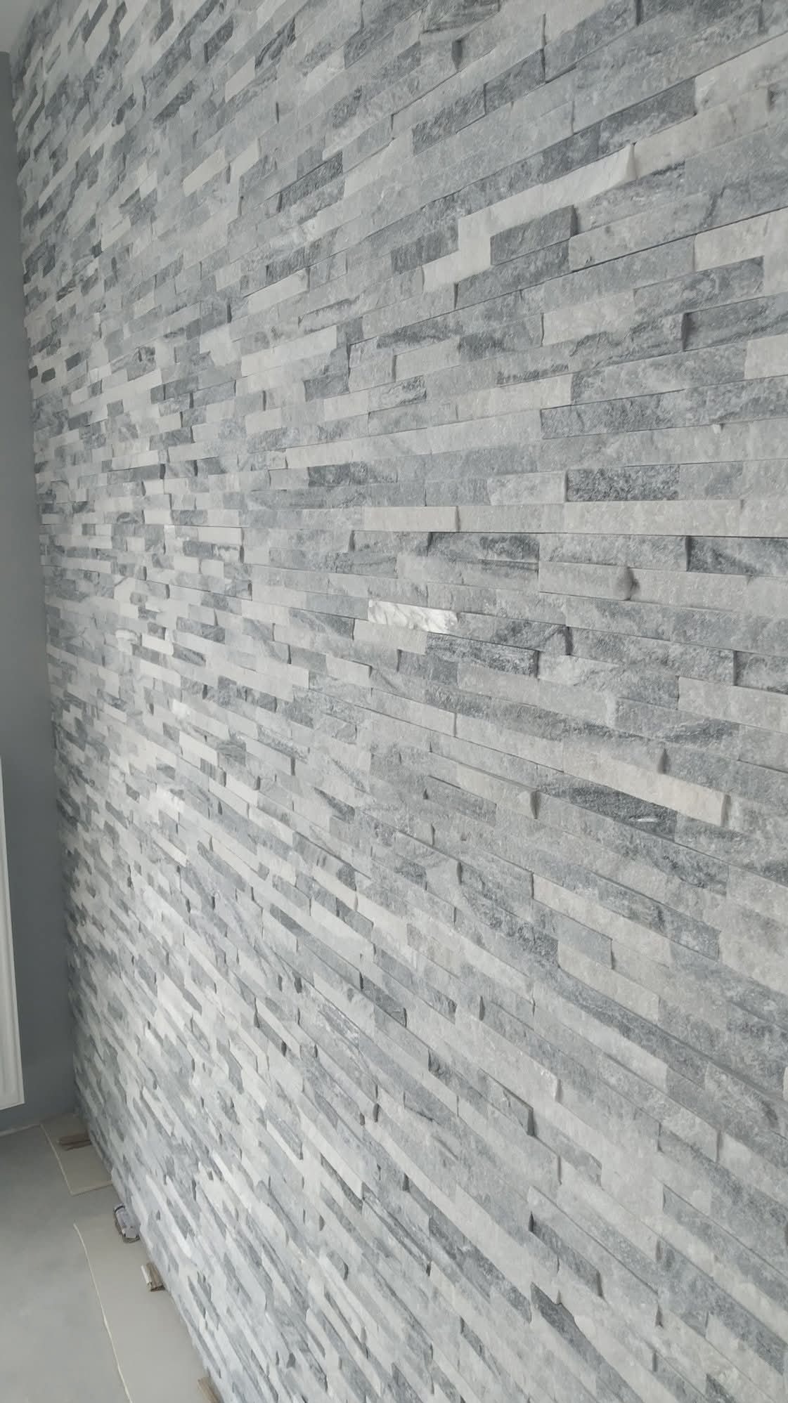 Ściana wykończona szarą, kamienną okładziną w wąskie, poziome pasy. Widok z boku, ukazujący fakturę i ułożenie elementów. Minimalistyczny design.