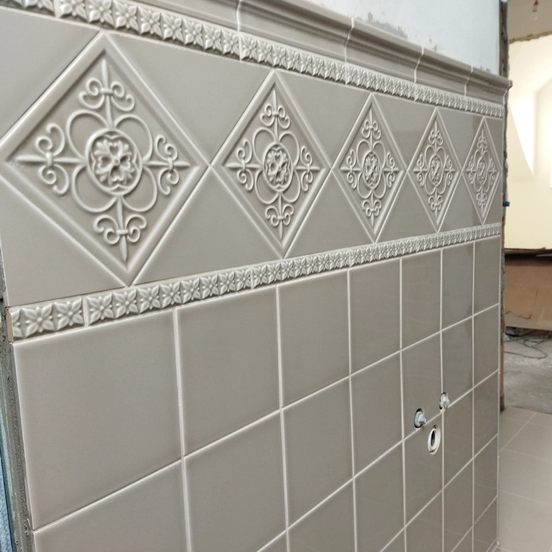 Ściana wyłożona szarymi, kwadratowymi płytkami z dekoracyjnym pasem z ornamentami kwiatowymi. Widoczne otwory na instalacje. Ukończony etap prac wykończeniowych.