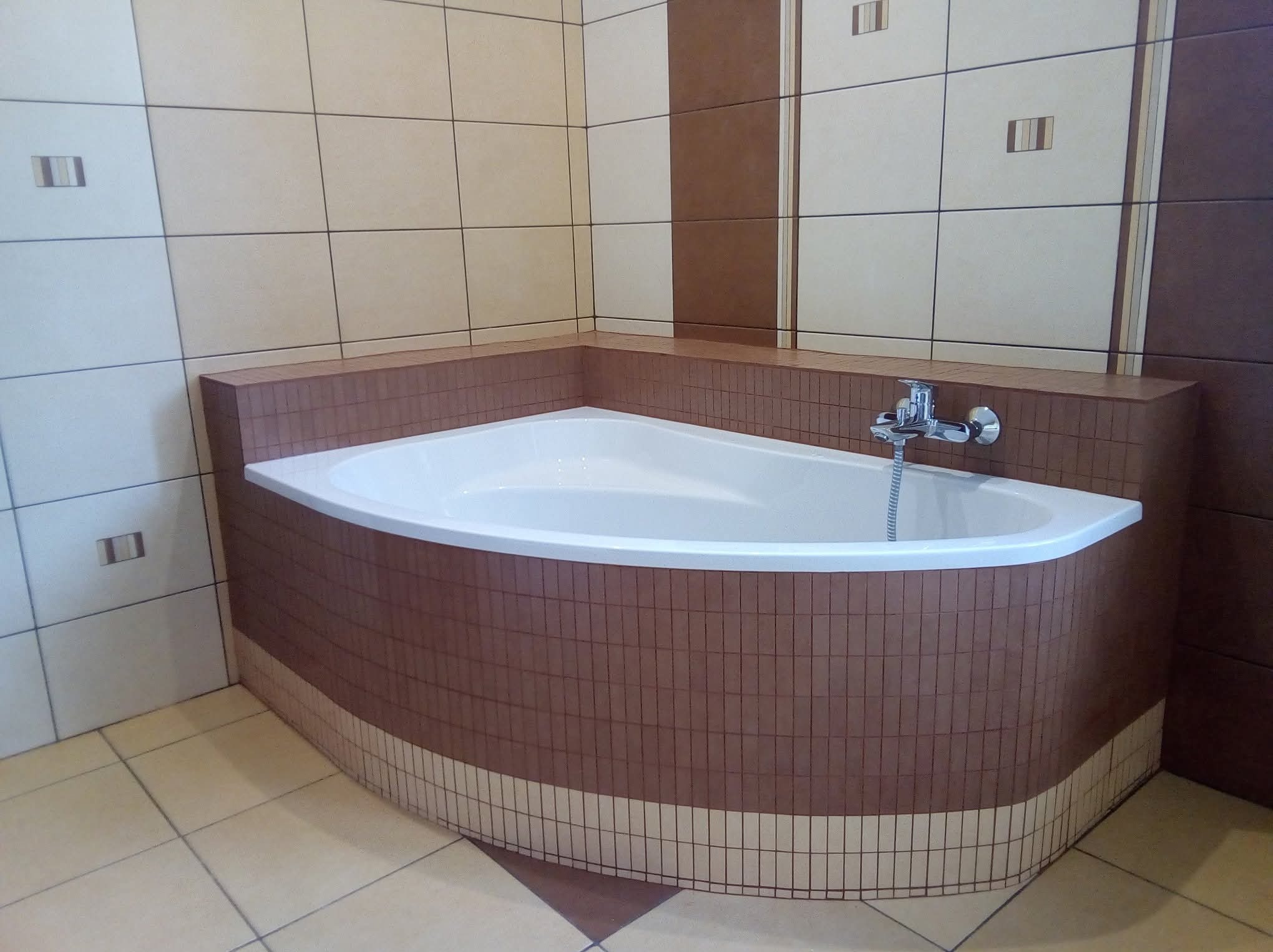 Narożna wanna obudowana brązowymi płytkami, z chromowaną baterią. Ściany wyłożone beżowo-białymi płytkami z dekoracyjnymi wstawkami. Podłoga z płytek w odcieniach beżu i brązu.