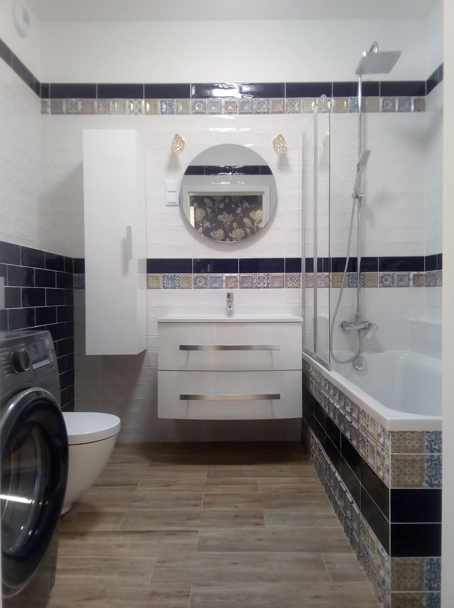 Wykończona łazienka z drewnopodobną podłogą, białą armaturą, dekoracyjnymi płytkami i okrągłym lustrem z podświetleniem. Widoczna pralka i wanna z parawanem.